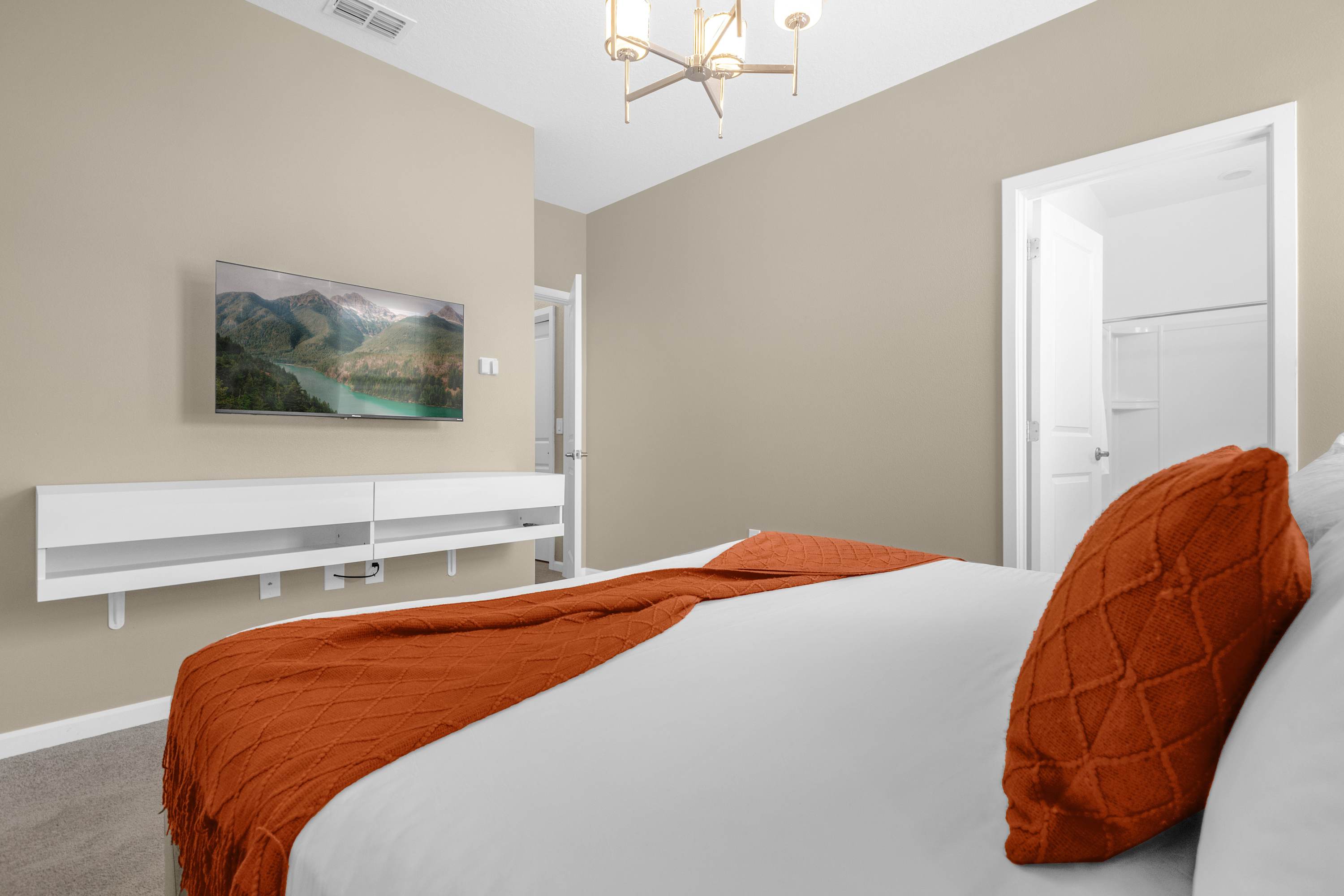 Modern bedroom with a large bed featuring an orange throw blanket and pillow, a wall-mounted TV displaying a nature scene, and a white floating media console against a light beige wall.