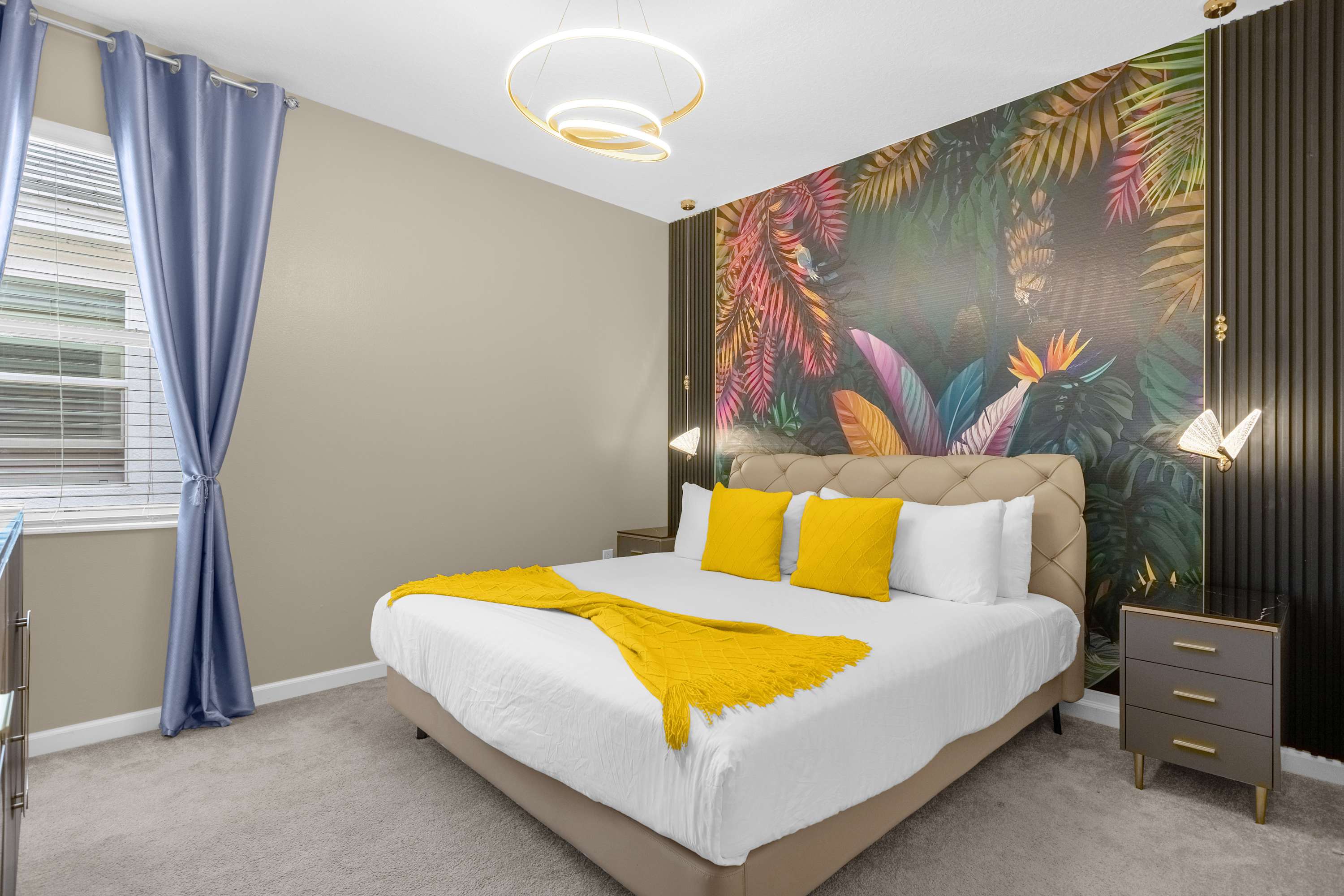 A vibrant bedroom featuring a king-sized bed with a beige upholstered headboard, bright yellow pillows, and a matching throw blanket. A striking tropical mural accentuates the wall behind the bed, flanked by dark fluted panels and unique hanging lights.