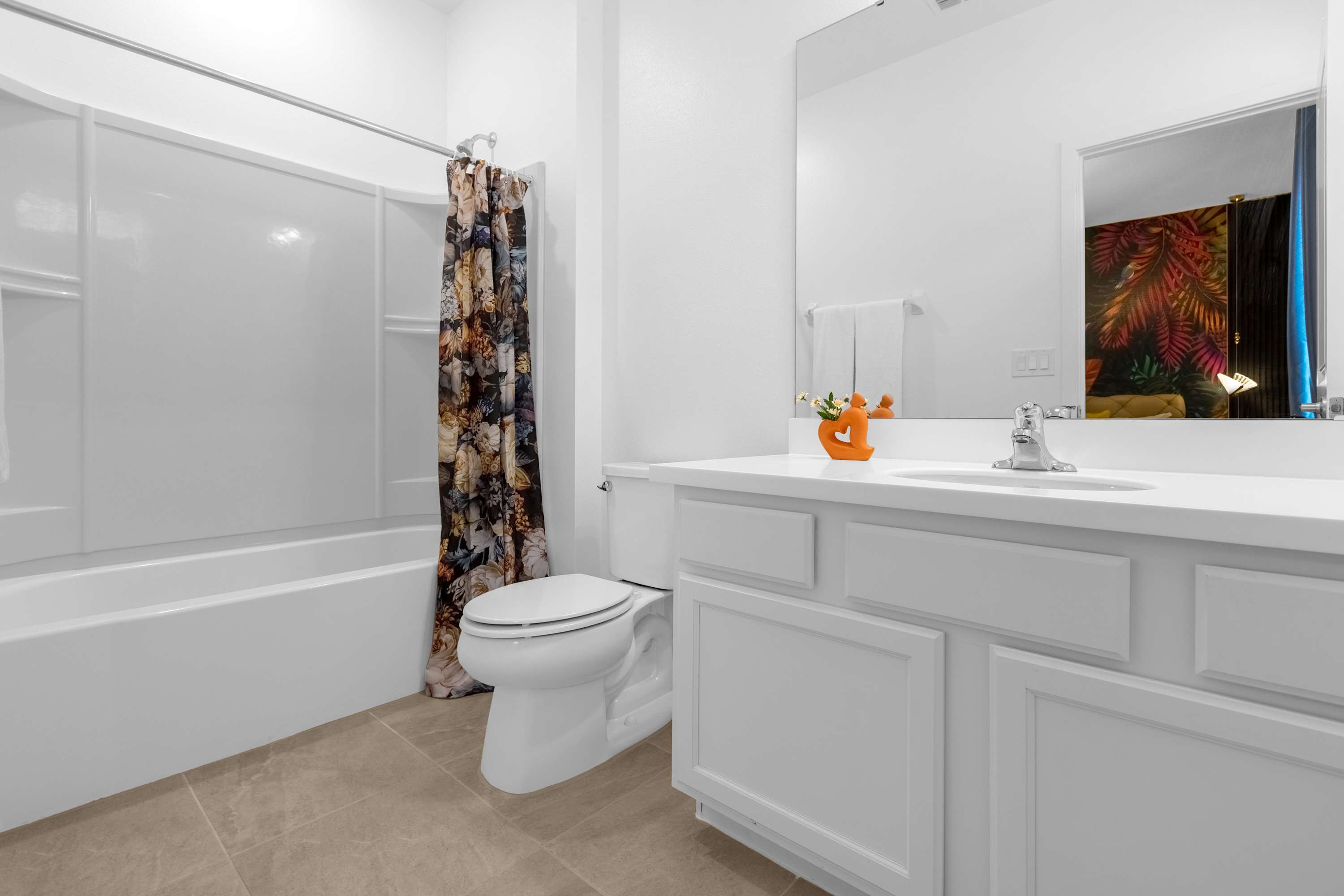 A clean, white bathroom featuring a single vanity with a white countertop and cabinets, a toilet, and a white tub/shower combo with a colorful, nature-themed shower curtain. A small plant and orange decor sit on the vanity.
