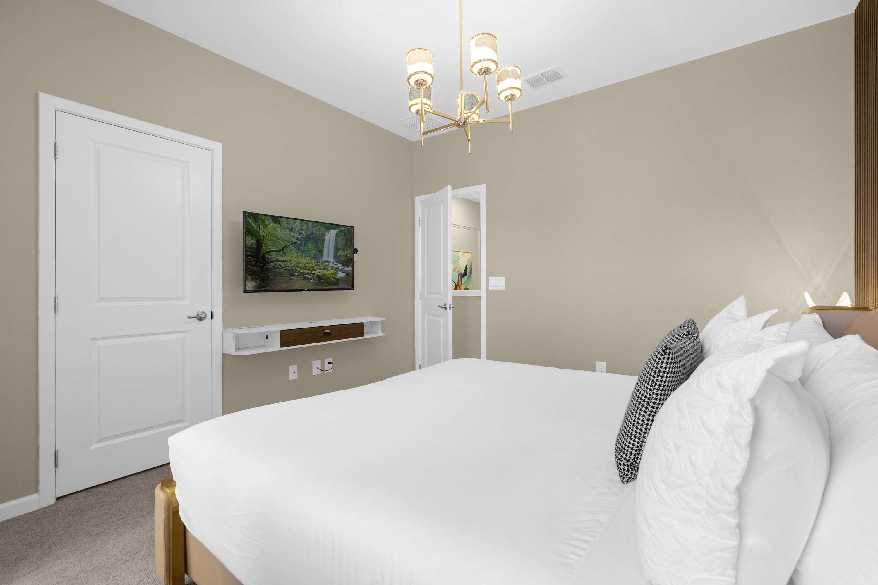 View of the contemporary bedroom showing the king bed, a wall-mounted TV above a floating shelf, and a doorway leading to another room, with light beige walls.