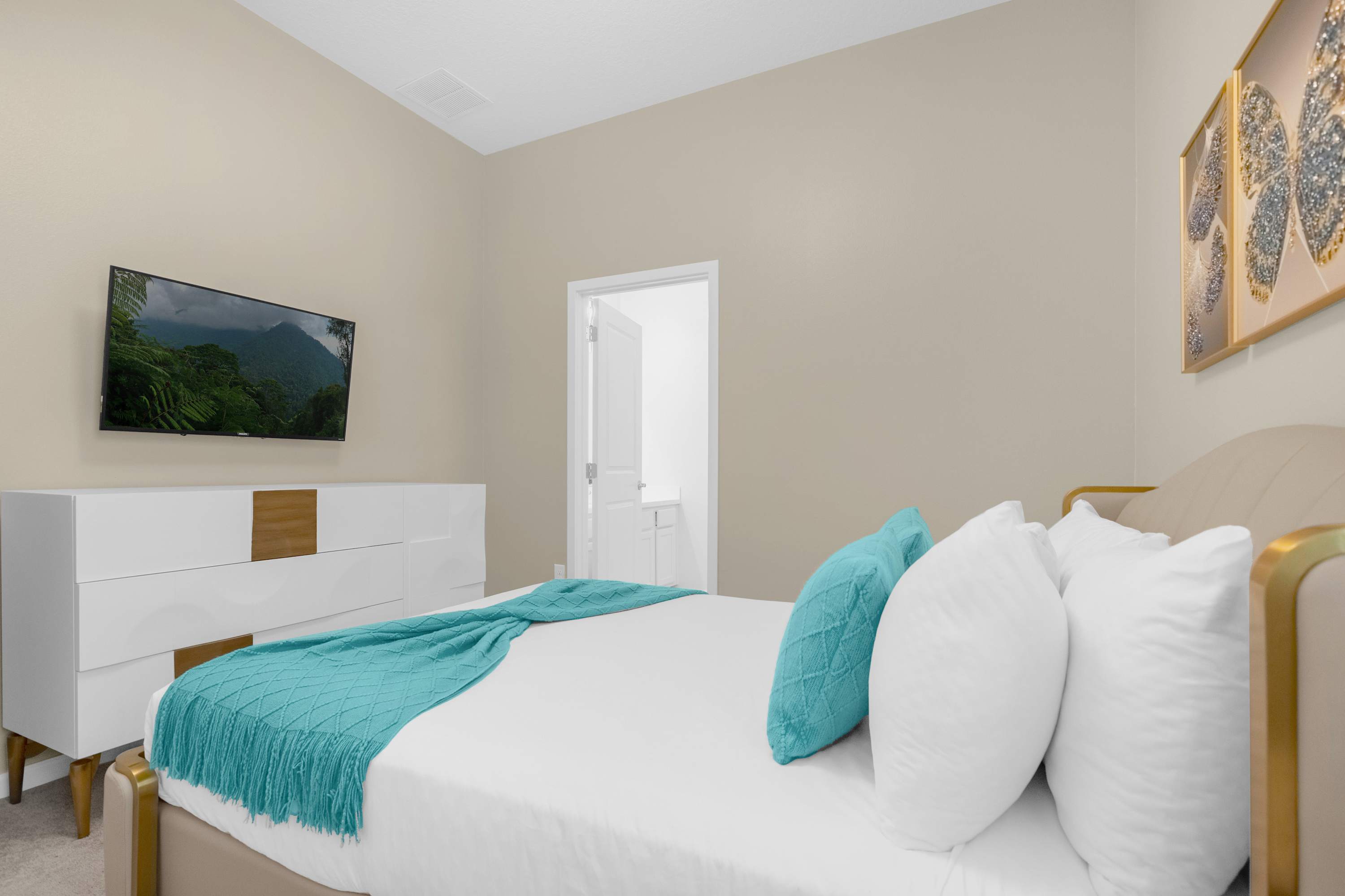 View of a cozy bedroom with a queen-sized bed dressed in white and teal, facing a wall-mounted flat-screen TV above a white and wood dresser. A doorway leads to an en-suite bathroom, and white closet doors are visible.