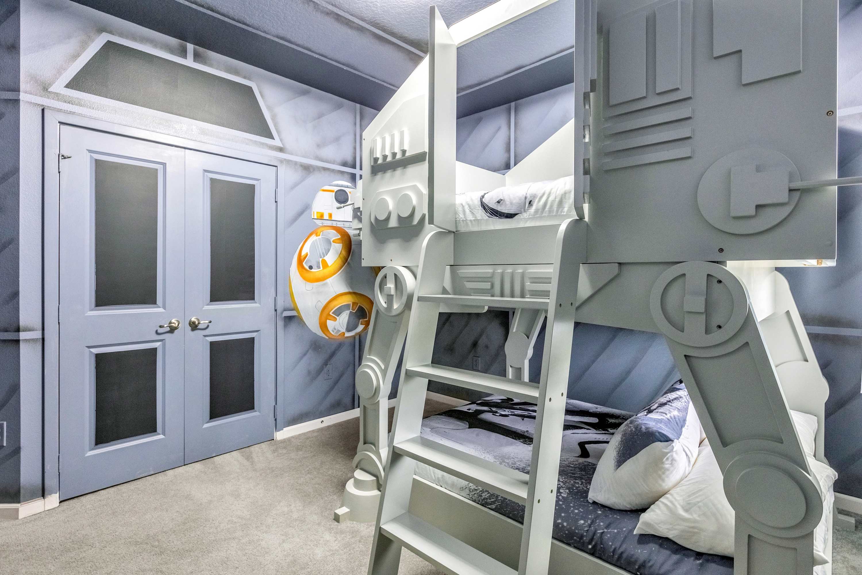 A Star Wars-themed bedroom featuring a grey AT-AT walker bunk bed with two sleeping areas, a BB-8 droid mounted on the wall, and grey paneled walls with a door designed to look like a spaceship hatch.