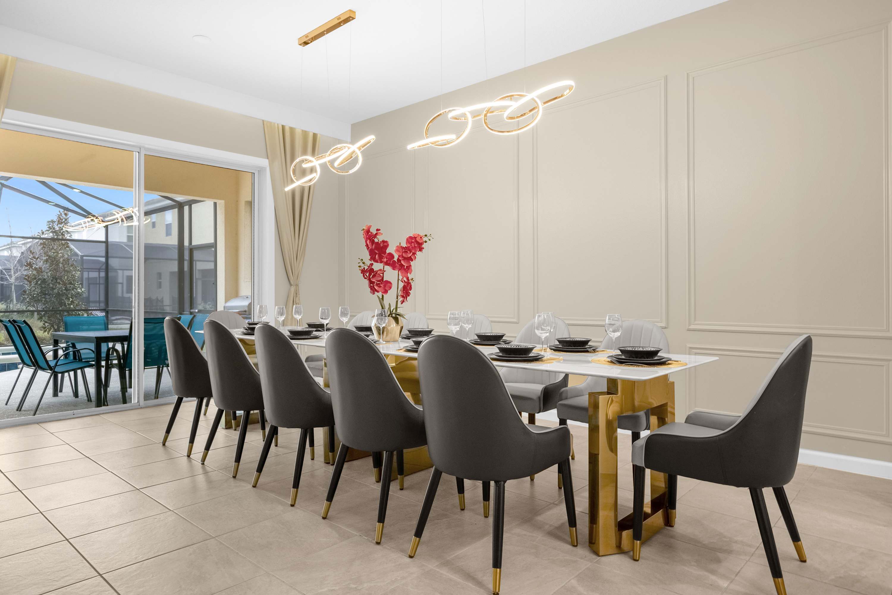 A spacious dining room with a long white dining table and eight dark grey upholstered chairs with gold accents. Modern, abstract gold light fixtures hang above the table, and a large sliding glass door opens to an outdoor patio area.