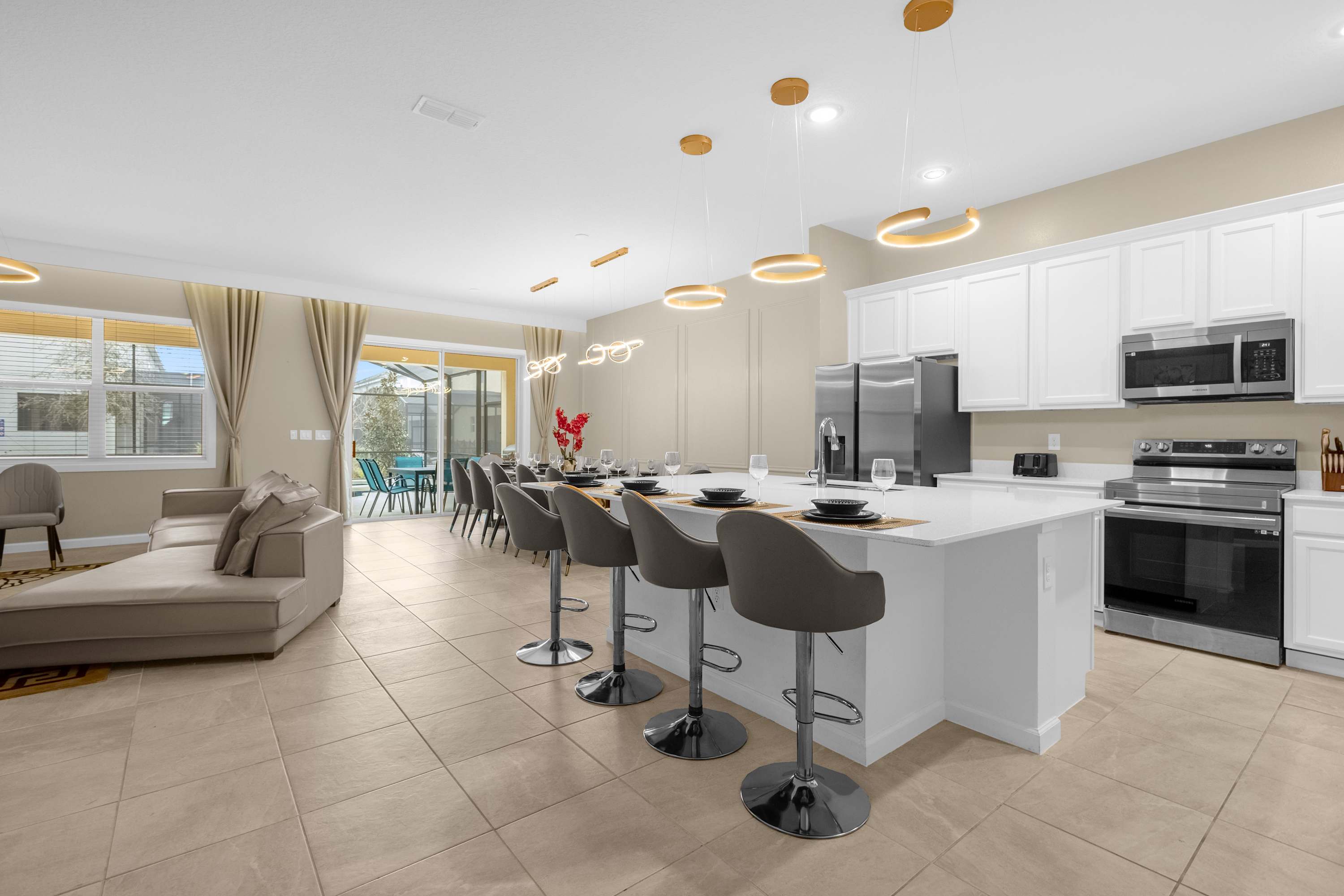 A wide view of an open-concept living space, showcasing a modern kitchen island with bar seating, a comfortable sectional sofa in the living area, and a formal dining table. The room features light tile flooring and contemporary lighting fixtures.