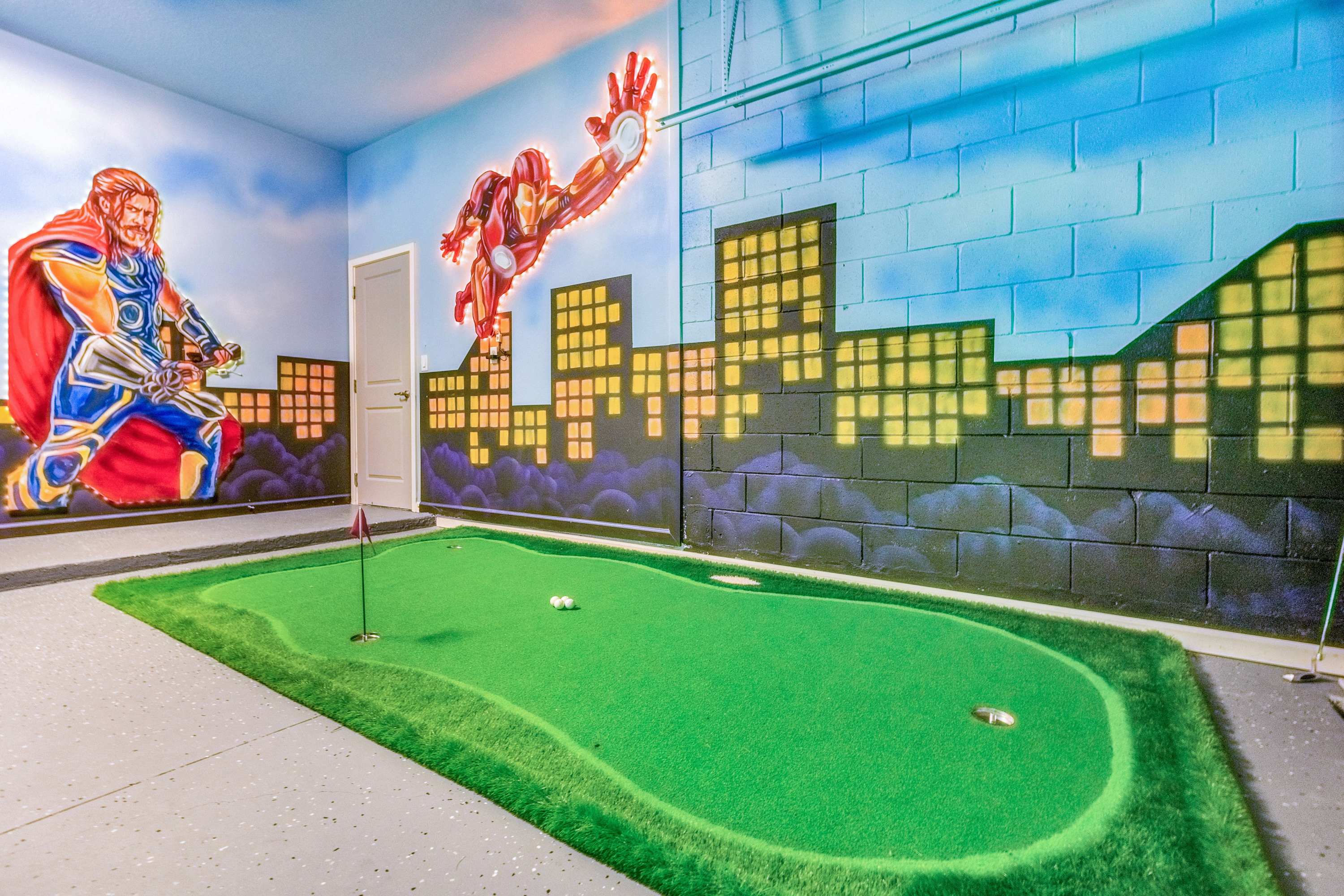 A close-up view of the putting green in the themed game room, featuring a flag and two golf balls. The walls are decorated with superhero murals and a cityscape design, creating an immersive play environment.