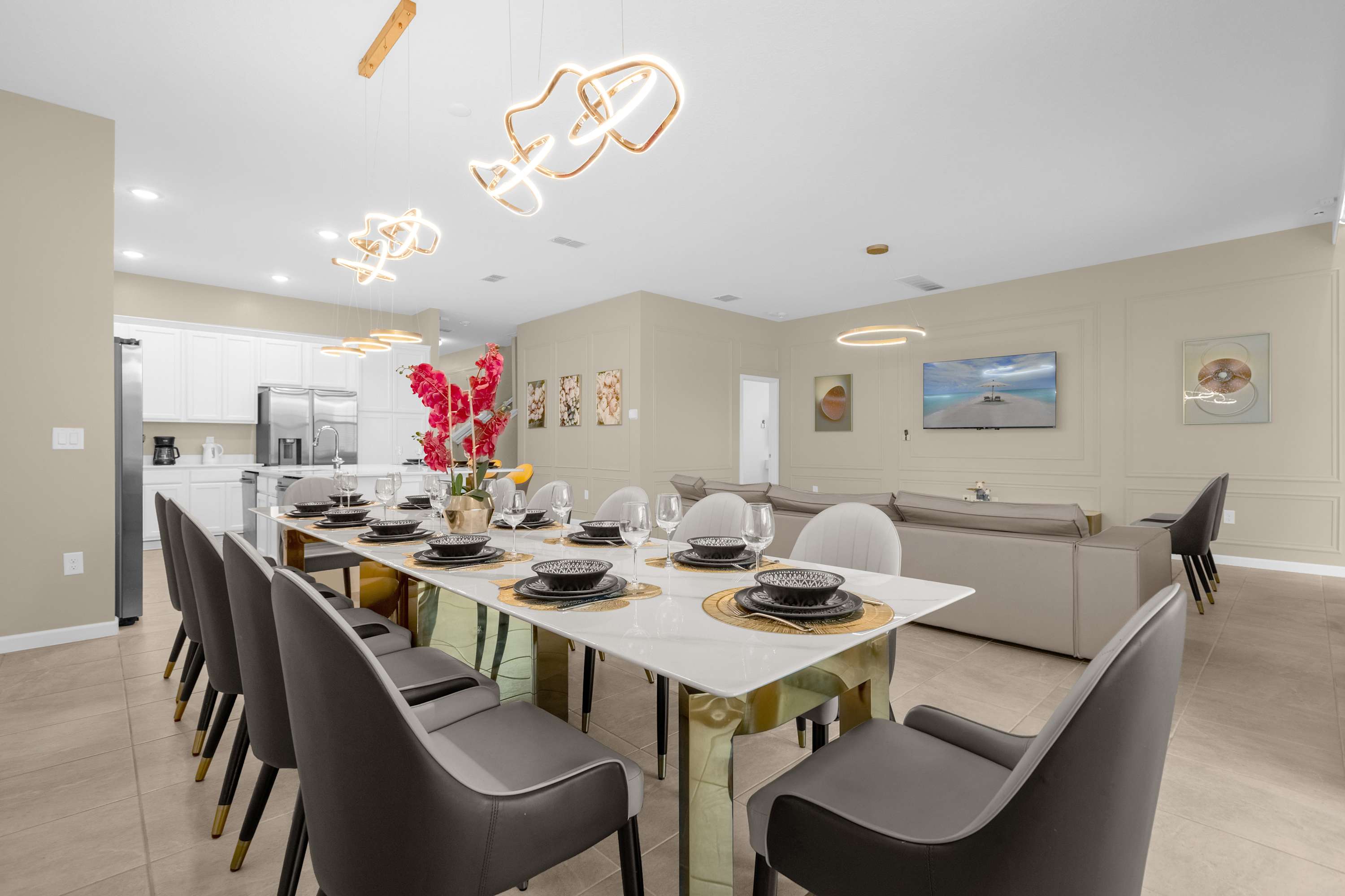 An open-concept dining area features a long white and gold dining table set for many guests, with modern gray chairs. The space flows into a contemporary kitchen with white cabinets and a living area with a large sectional sofa, all under unique gold-toned chandeliers.