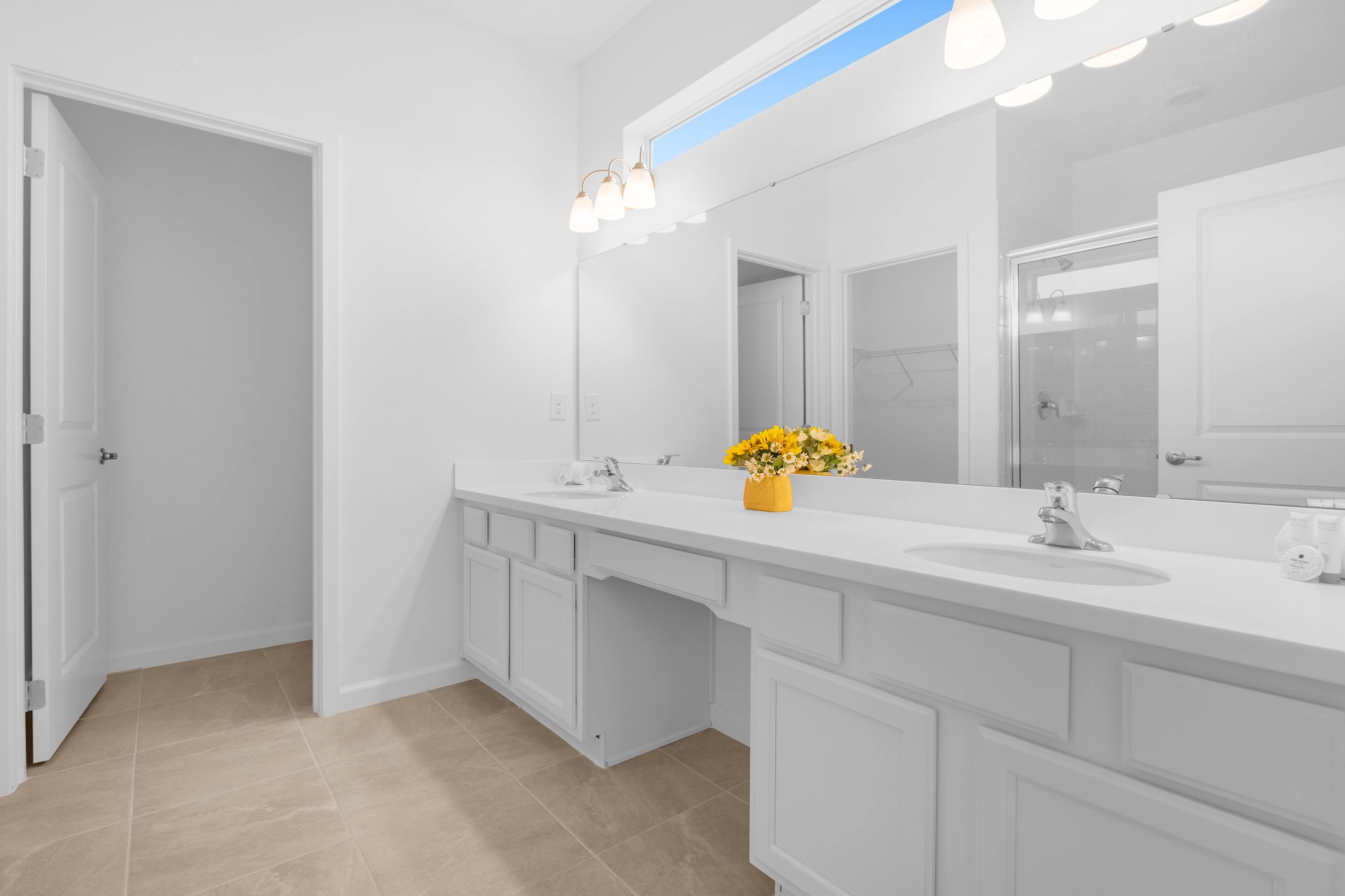 Spacious and bright white bathroom featuring a long double vanity with two sinks, a large mirror, and light-colored tiled flooring, with a yellow floral arrangement on the counter.