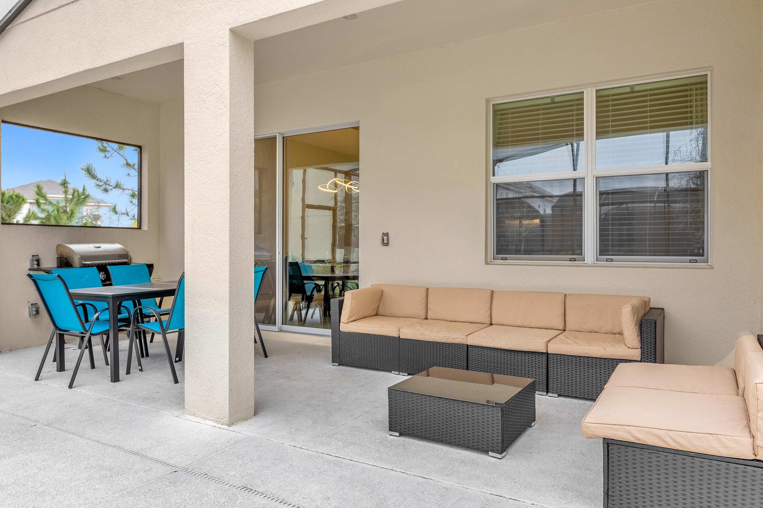 A covered outdoor patio area features a long beige sectional sofa with a dark coffee table, and a separate dining area with a grill and blue chairs. Large windows and a sliding glass door connect the patio to the interior of the house.