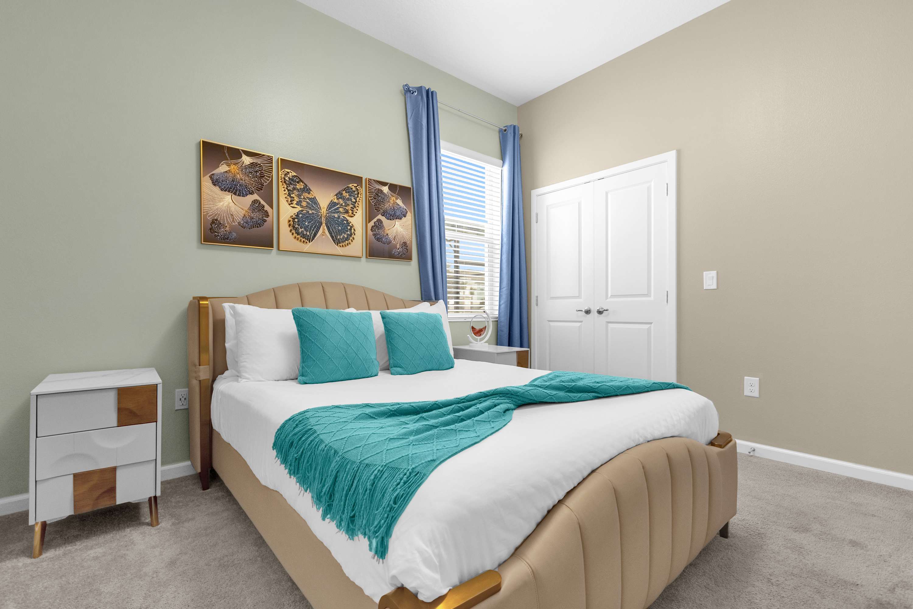 A serene bedroom featuring a queen-sized bed with a beige upholstered headboard, white linens, and teal decorative pillows and a matching throw blanket. Butterfly artwork adorns the wall above the bed, and a window with blue curtains provides natural light.