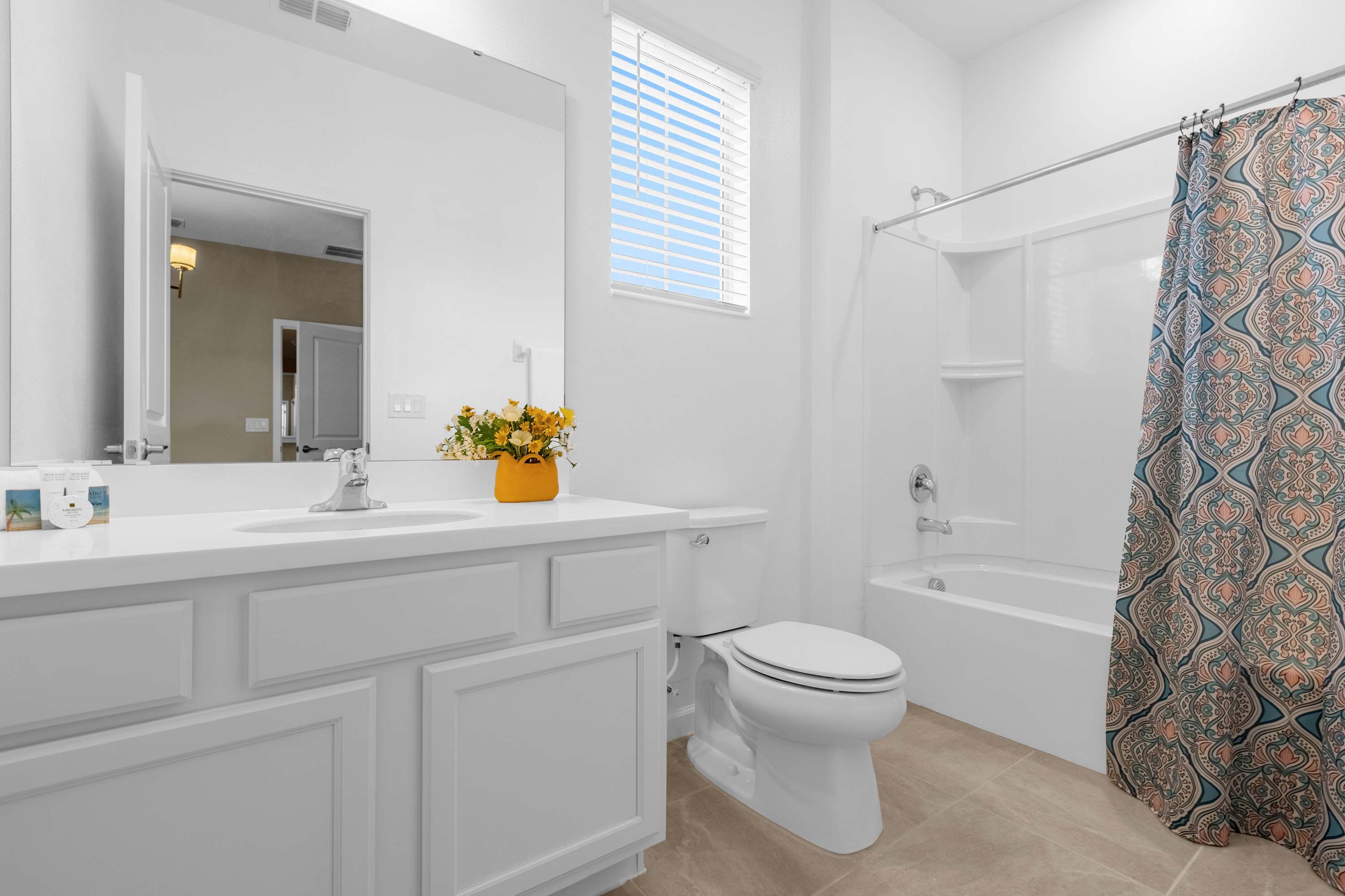 Clean white bathroom featuring a single vanity with a sink, a toilet, and a shower-tub combo with a colorful patterned curtain, illuminated by natural light from a window.