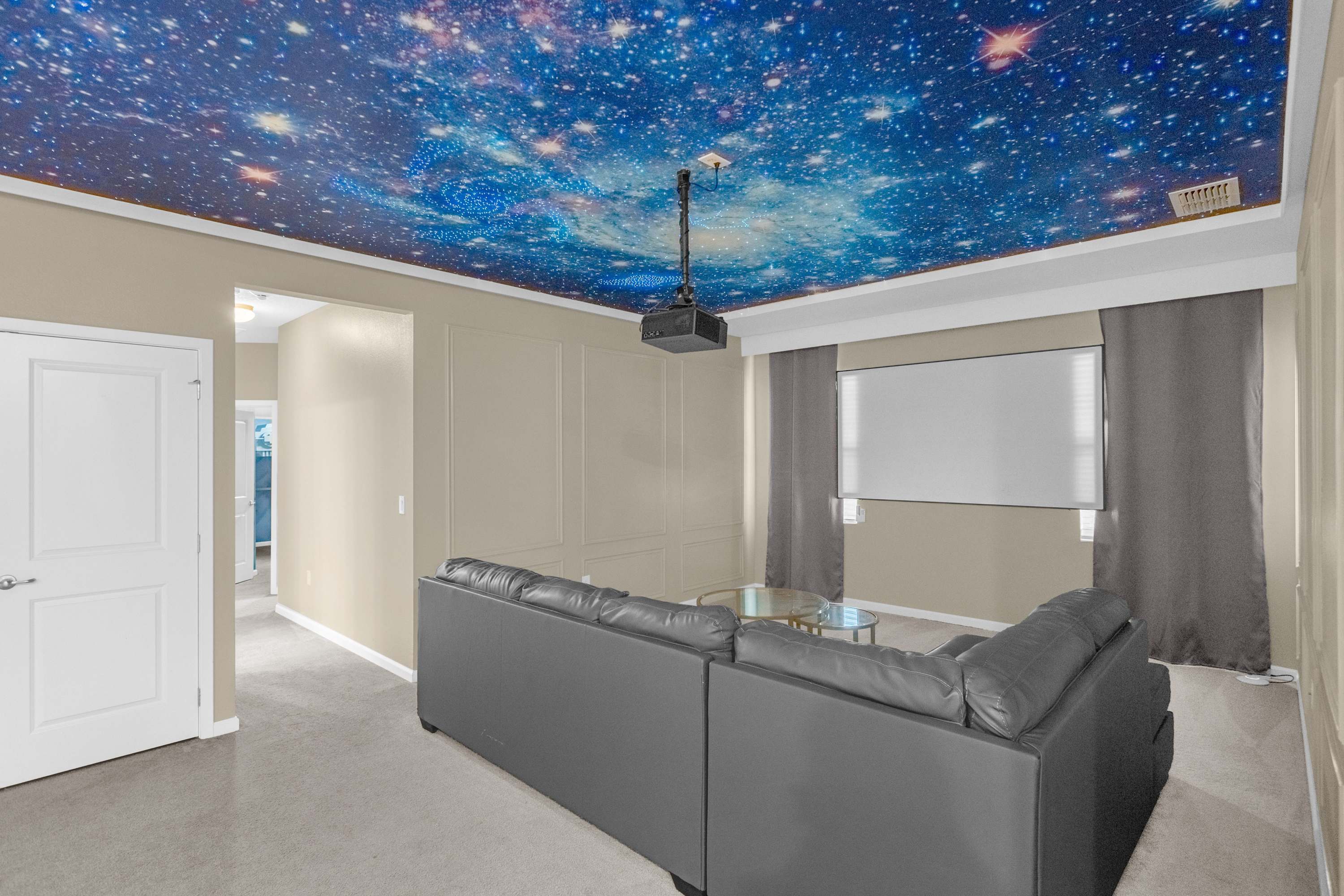 A wide shot of the media room, highlighting the large projector screen and the comfortable grey sectional sofa. The striking galaxy-themed ceiling mural is prominent, and the room is equipped with dark curtains for optimal viewing.