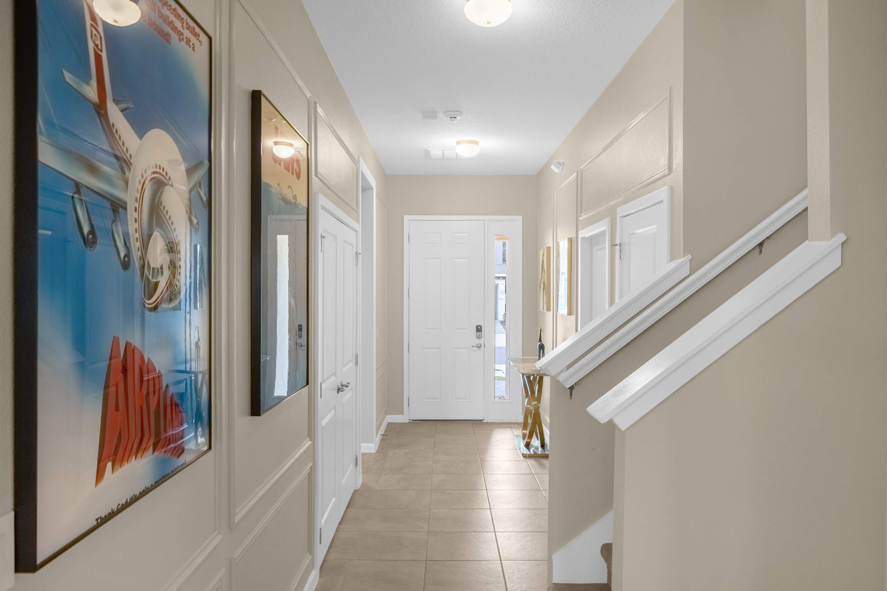 Bright entryway featuring a white front door with glass inserts, light beige walls with wainscoting, tiled flooring, and a staircase leading upstairs, adorned with framed artwork on the left.