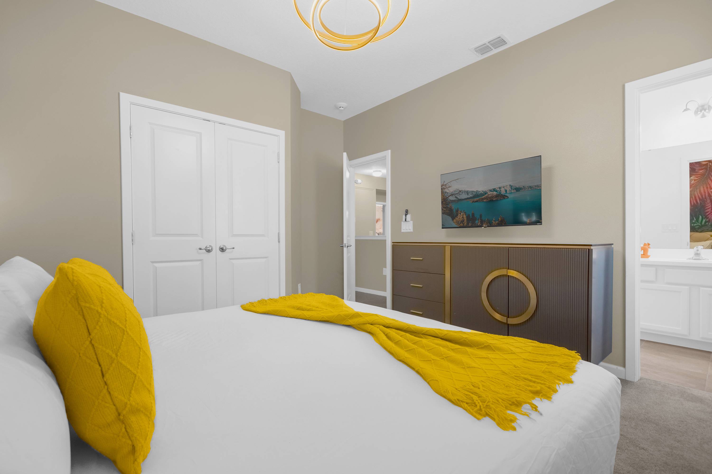 View of a modern bedroom with a king-sized bed dressed in white linens and yellow accents, facing a wall-mounted flat-screen TV above a dark wood dresser. White bi-fold closet doors and a doorway leading to a bathroom are visible.