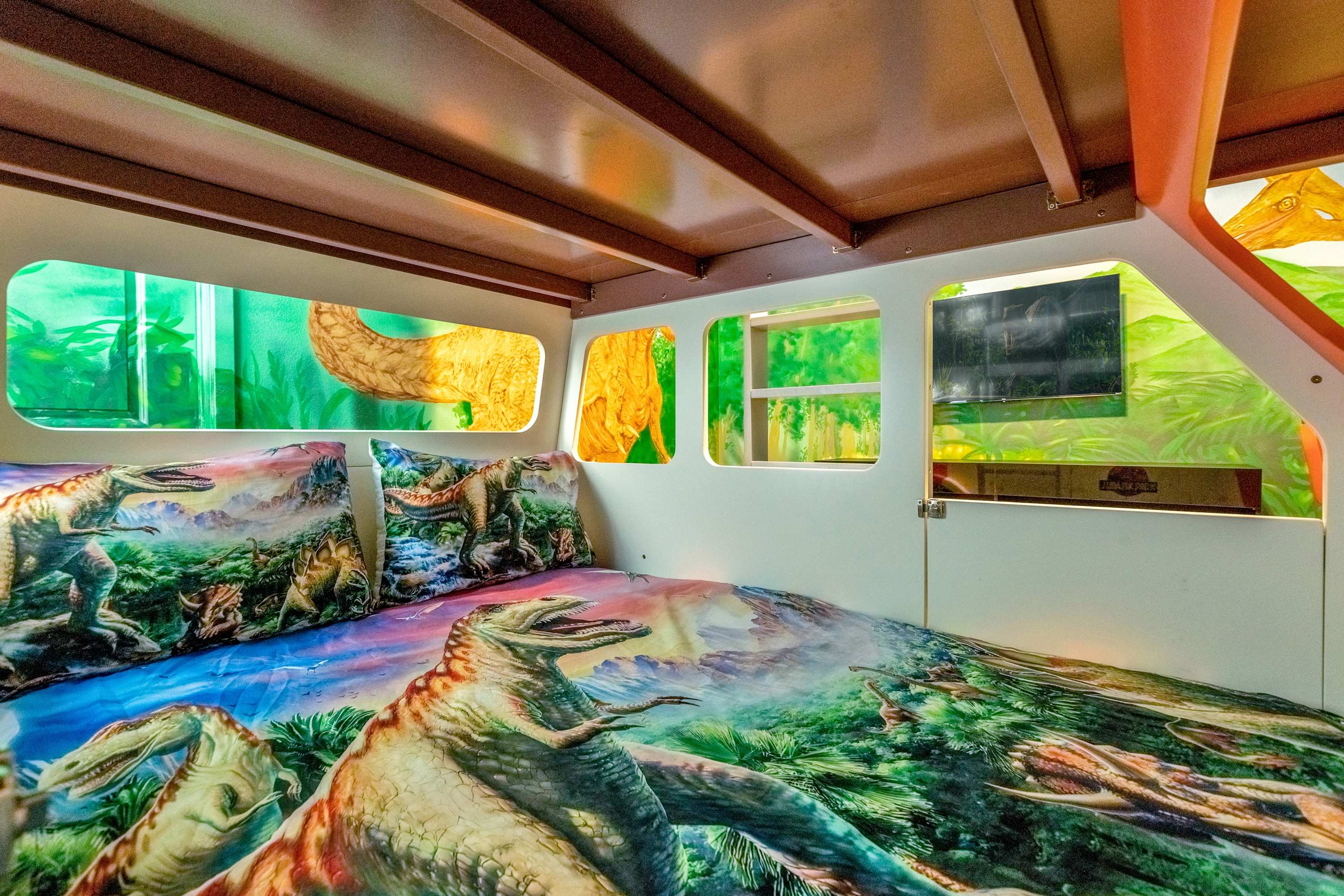 An interior view of the lower bunk bed in the Jurassic Park-themed room, showing dinosaur-themed bedding, a small wall-mounted TV, and windows cut into the vehicle structure.