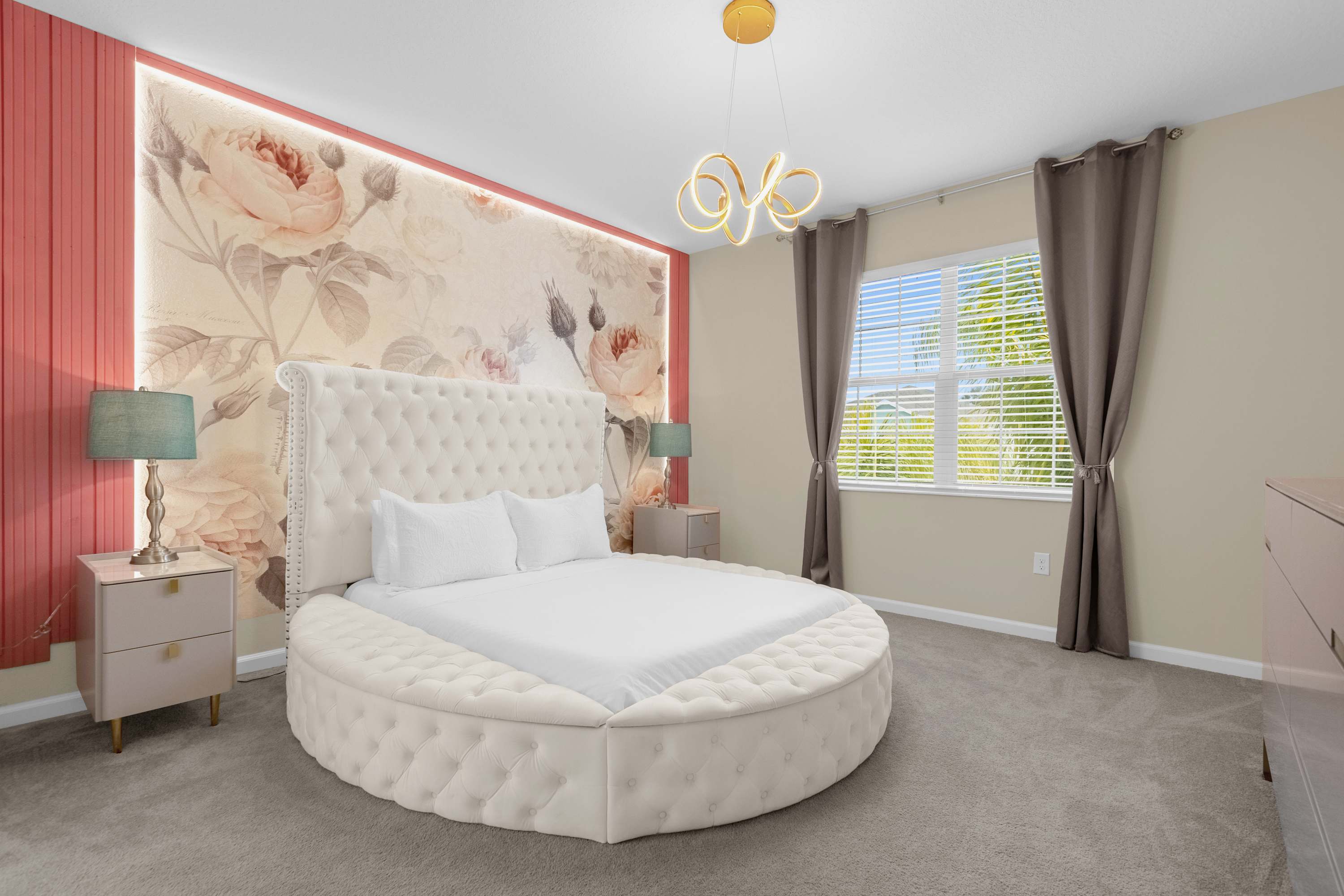 Elegant bedroom with a large tufted round bed, a feature wall adorned with large floral wallpaper, and a window with dark grey curtains, illuminated by a modern gold chandelier.