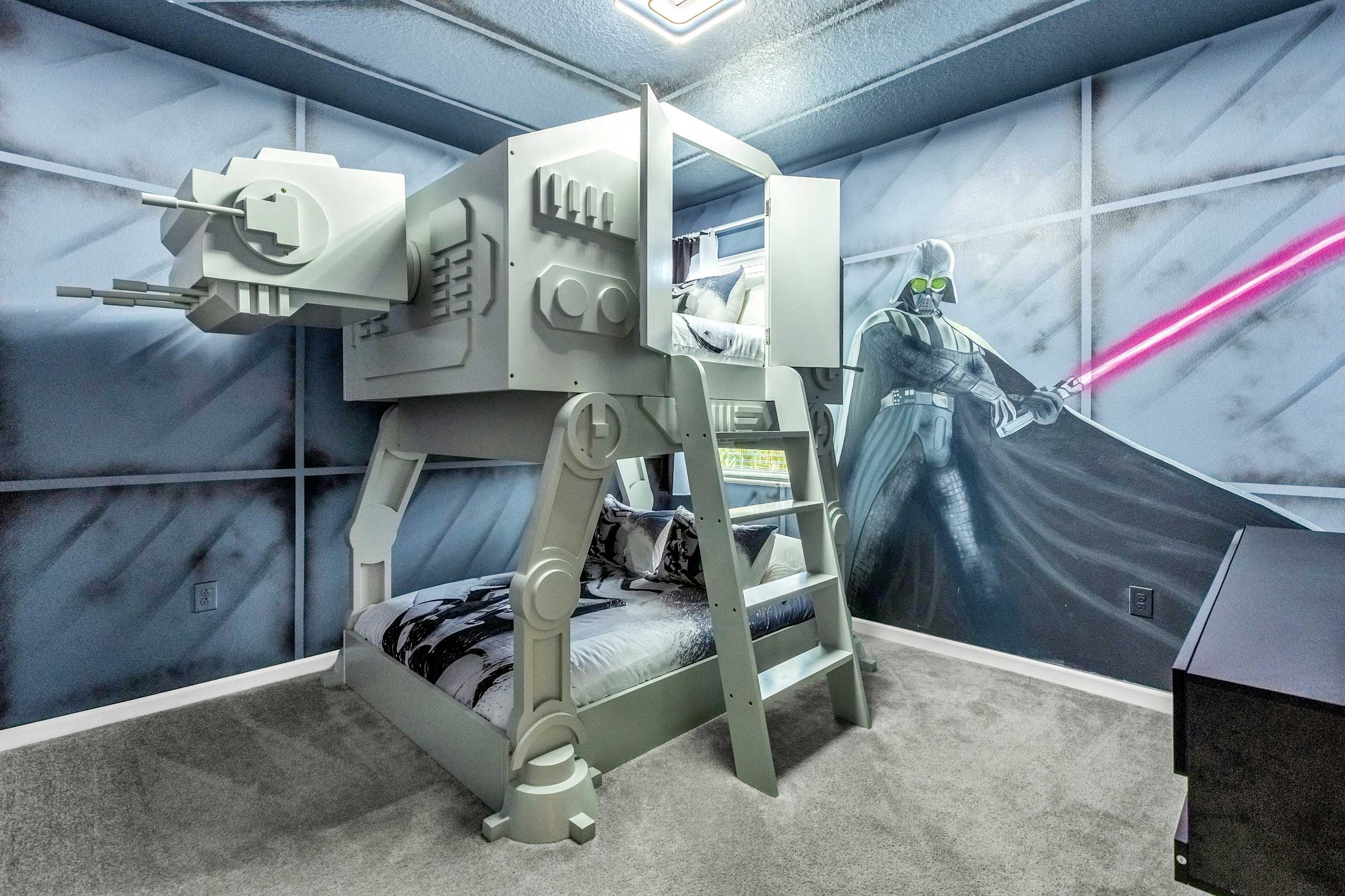 A Star Wars-themed bedroom featuring a large, grey AT-AT walker bunk bed with a lower bed and an upper sleeping area accessible by a ladder. The walls are painted with a grey, metallic panel design and a mural of Darth Vader wielding a pink lightsaber.