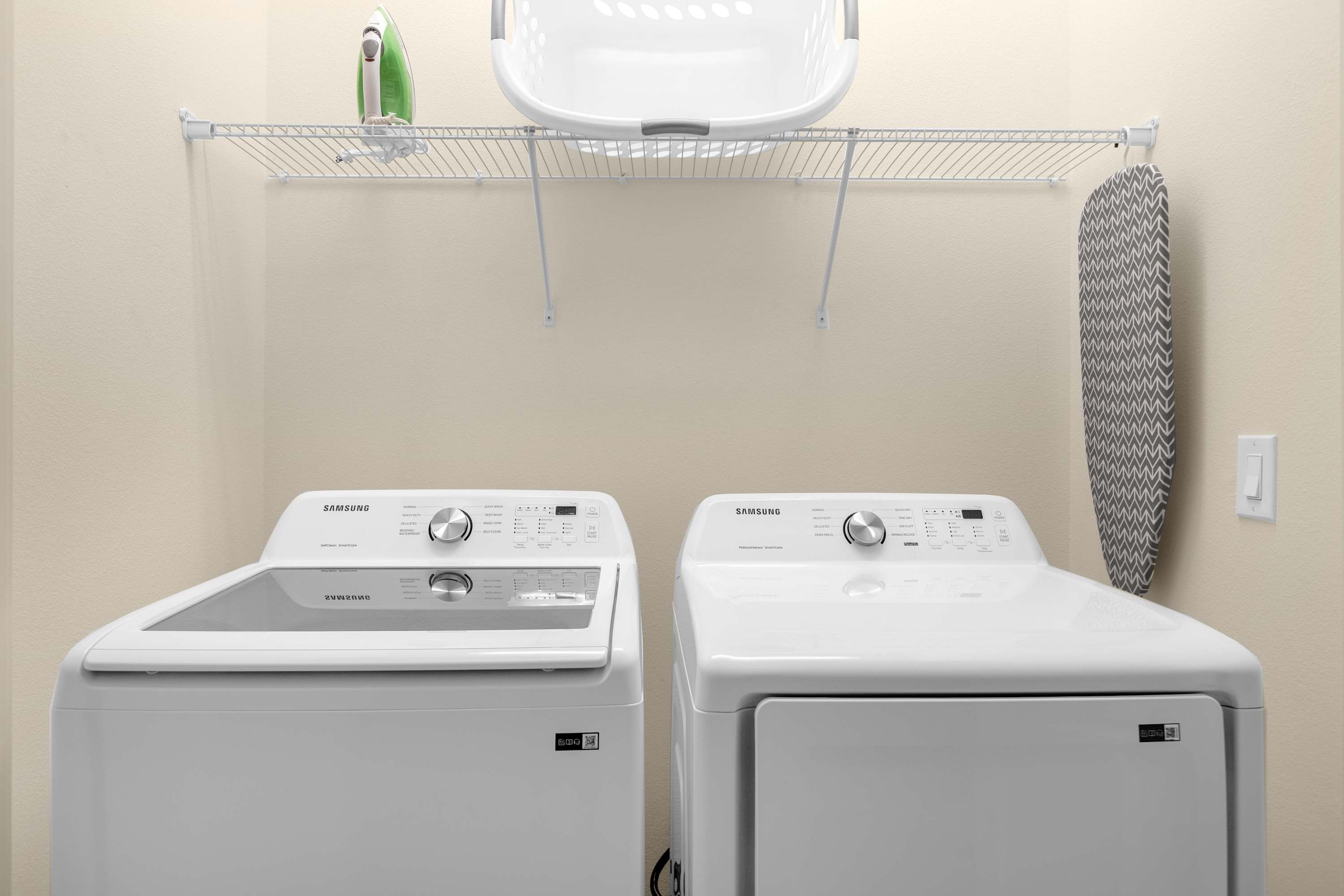 Functional laundry room equipped with a white Samsung top-load washer and front-load dryer, a wire shelf with a laundry basket, and an ironing board hanging on the wall.