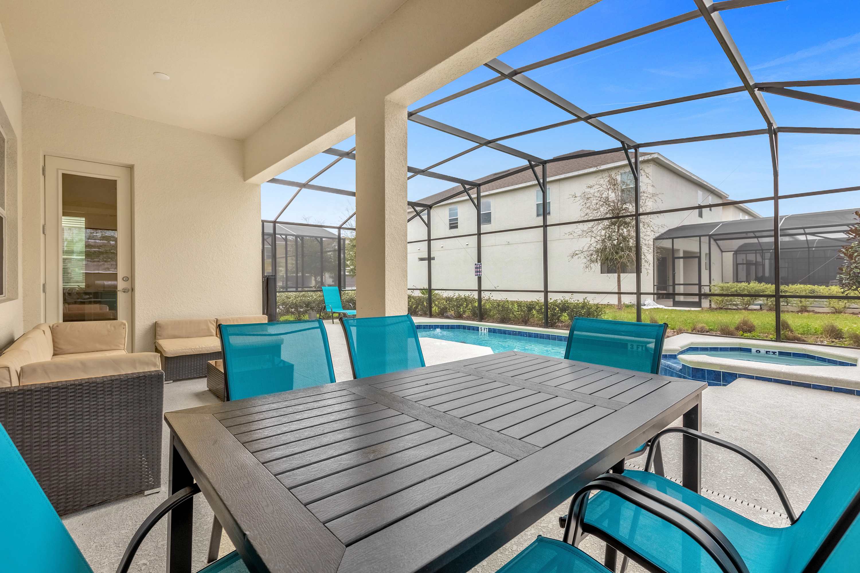 A covered outdoor patio area within a screened enclosure features a dark dining table with bright blue chairs and a separate seating area with a beige sectional sofa. The pool is visible just beyond the patio, and a glass door leads indoors.