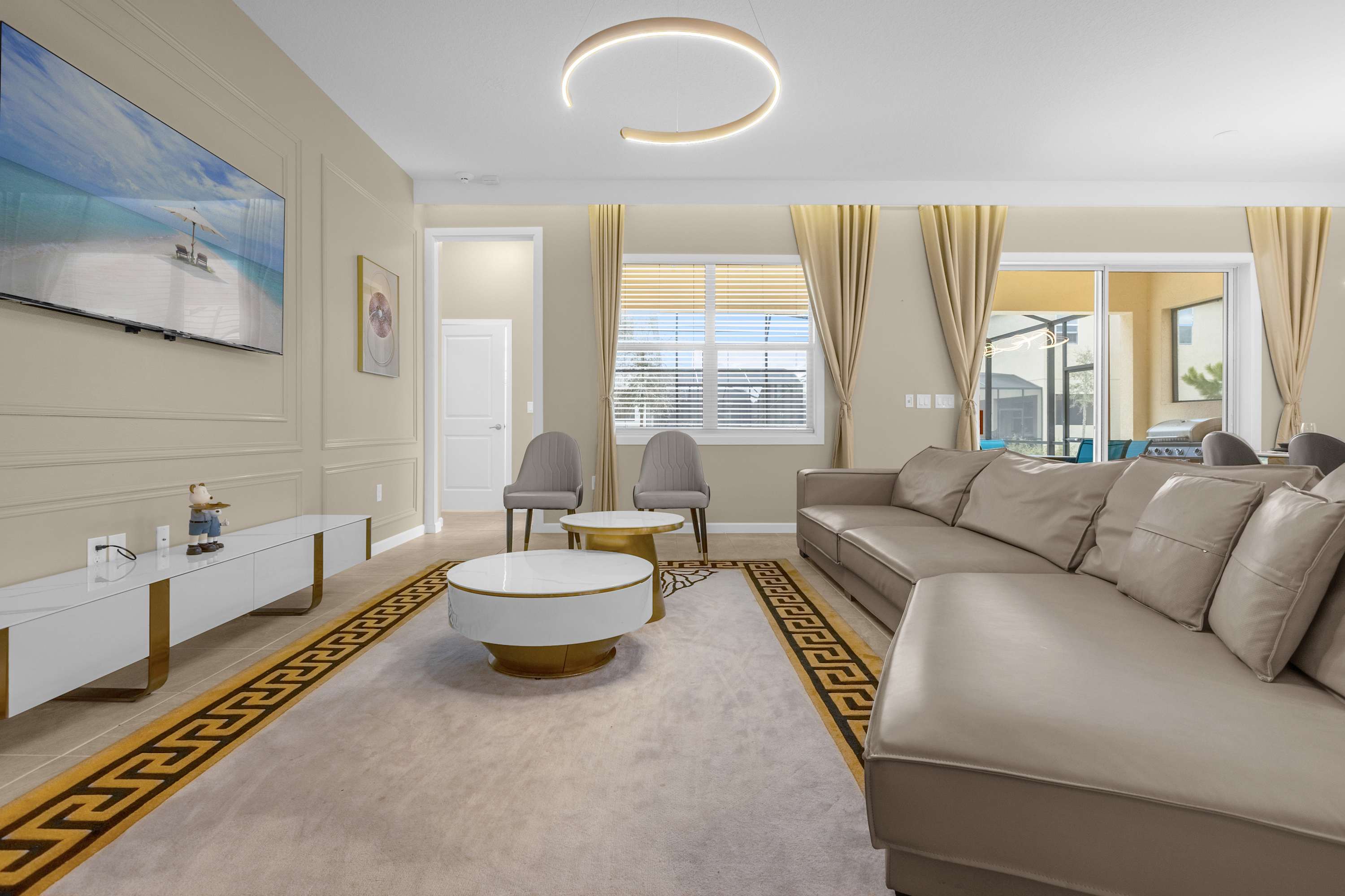 A modern living room featuring a large, light beige sectional sofa, two round coffee tables, and a wall-mounted TV. The room has light-colored walls with decorative paneling, a patterned rug, and a unique circular ceiling light fixture.