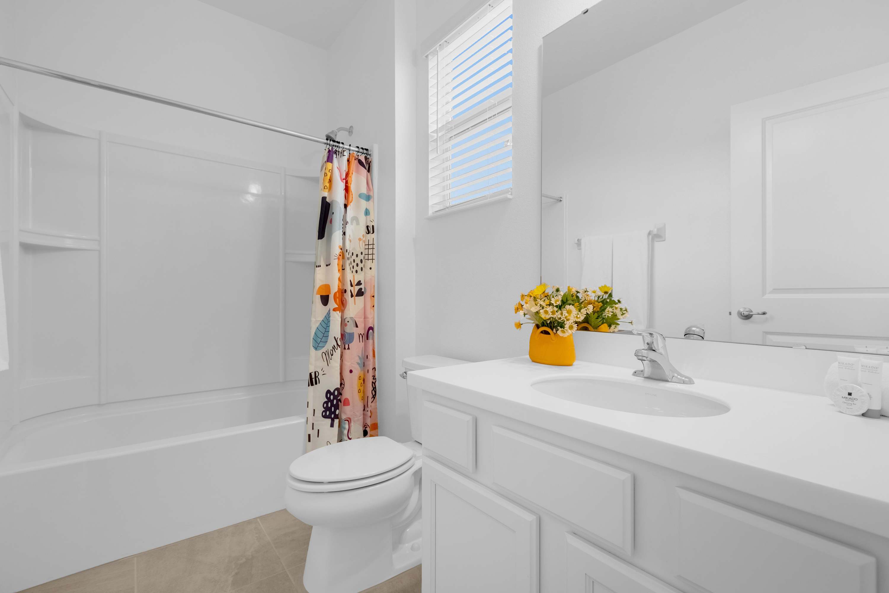 White bathroom with a single vanity, toilet, and a shower-tub combo featuring a vibrant, patterned shower curtain, illuminated by natural light from a window with blinds.