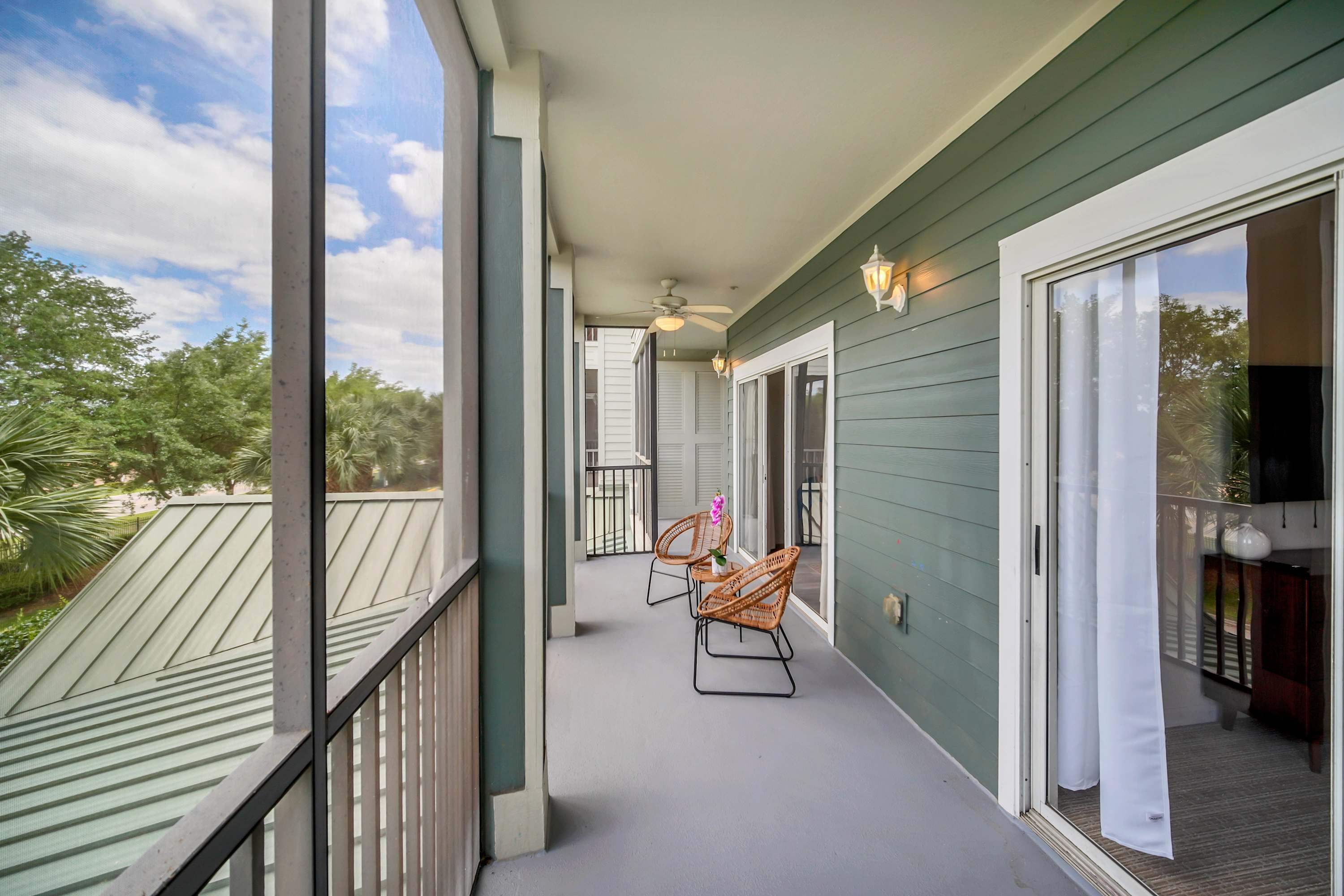 Long screened balcony with grey flooring, featuring two wicker chairs with black frames, offering views of the surrounding trees and other buildings.