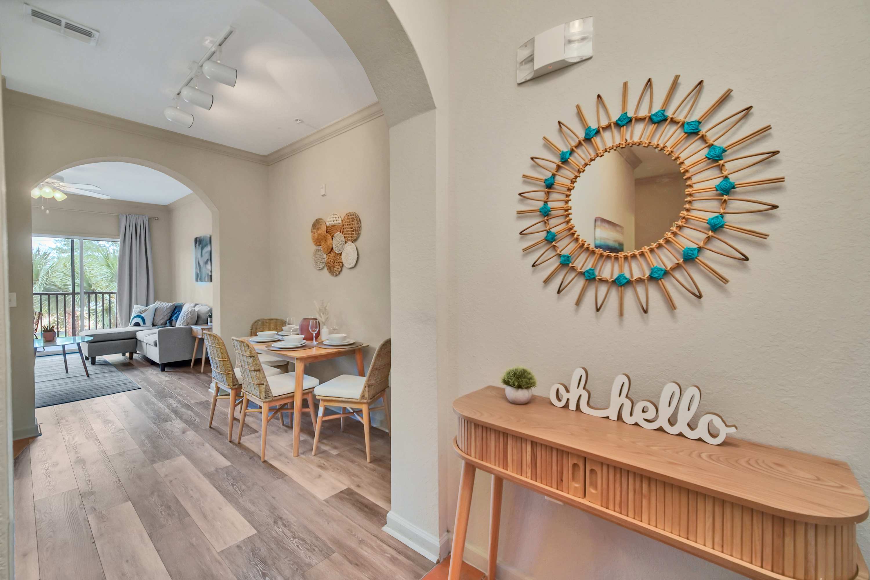 A bright entryway features a wooden console table with 'oh hello' decor and a round mirror with a rattan frame and blue accents, leading into an open-concept dining and living area with light wood flooring.