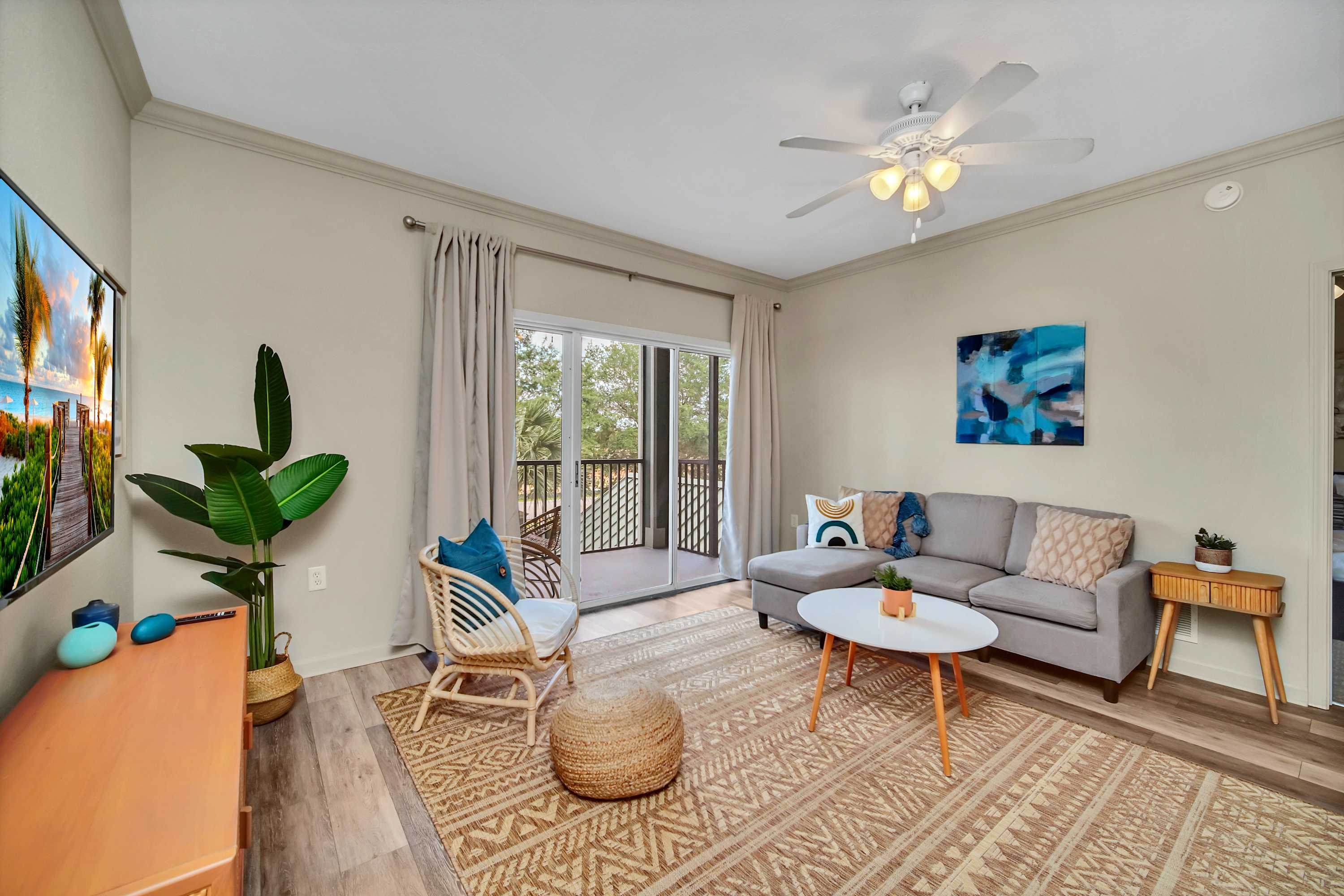 A slightly different angle of the living room, showing the light gray sectional sofa, white coffee table, and rattan chair on a patterned jute rug. The sliding glass doors lead to the balcony, and a ceiling fan is overhead.