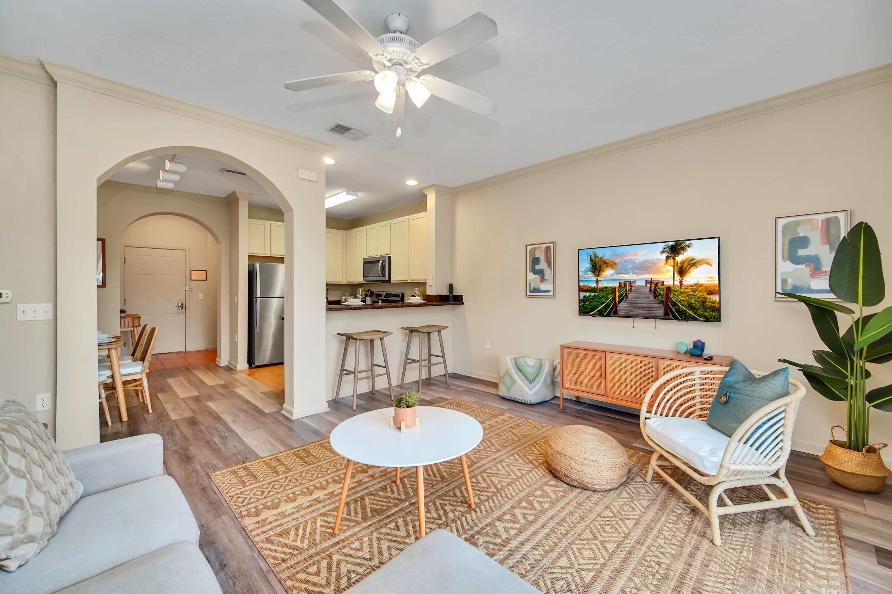 An open-concept living area flows into a dining space and kitchen, featuring a light gray sectional sofa, a white coffee table, and a rattan chair on a patterned jute rug. A breakfast bar with stools separates the living room from the kitchen.