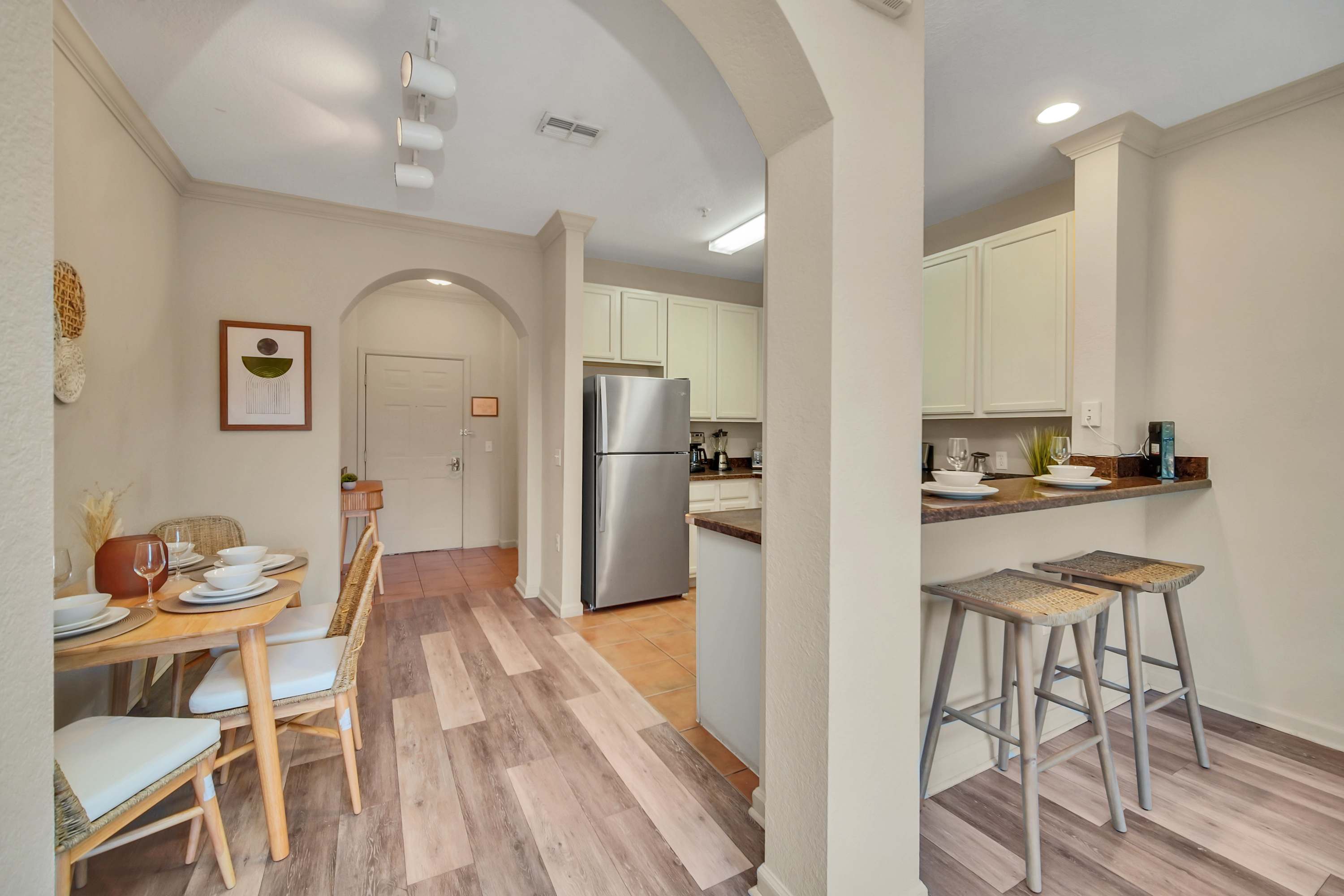 An open-concept space connects the dining area with a light wood table and woven chairs to the kitchen, which features a breakfast bar with two stools and stainless steel appliances, all under an arched entryway.