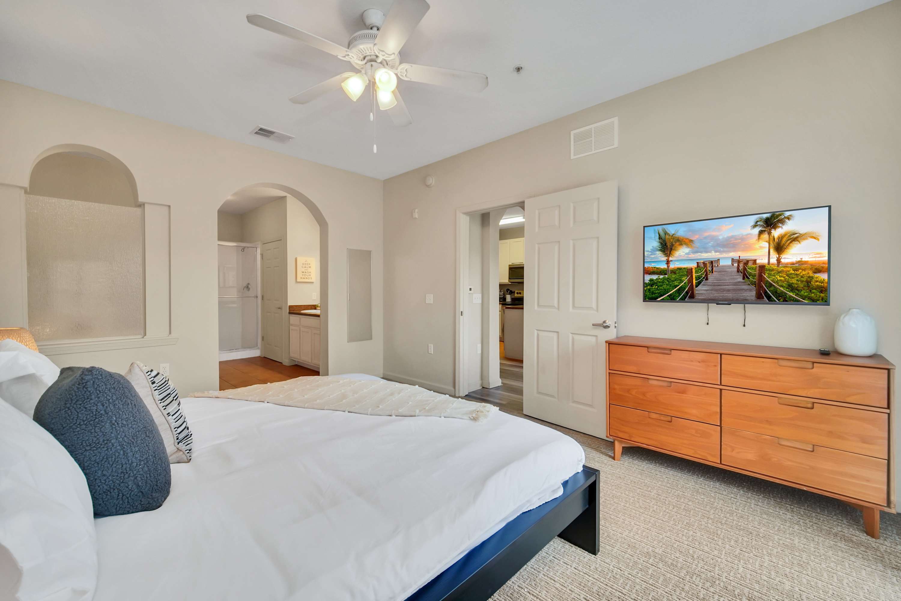 A cozy bedroom features a bed with white linens and blue accent pillows, a wooden dresser with a flat-screen TV mounted above it, and a ceiling fan, with an archway leading to an en-suite bathroom.