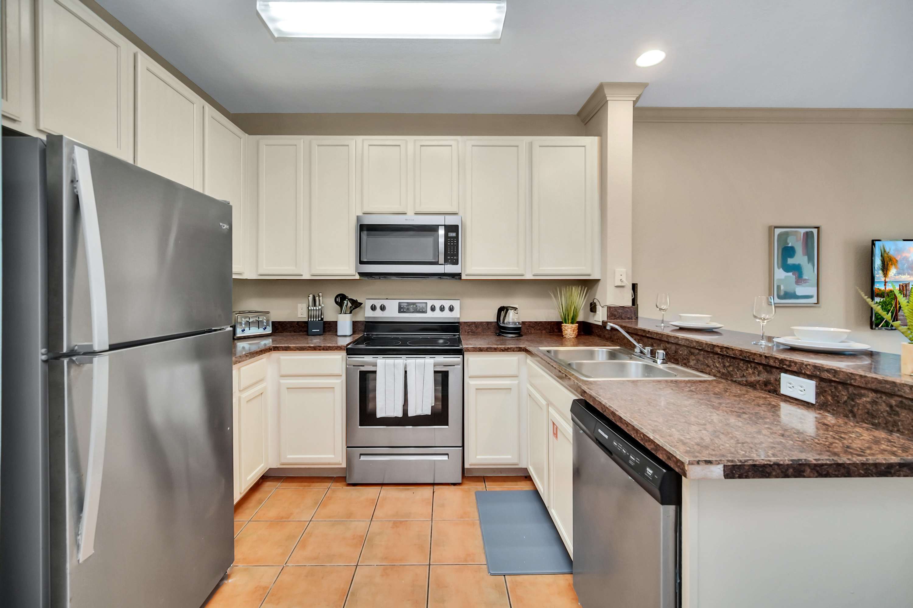 A spacious kitchen boasts stainless steel appliances, including a refrigerator, stove, and microwave, complemented by light cream cabinetry and dark granite-look countertops, with terracotta-colored tile flooring.