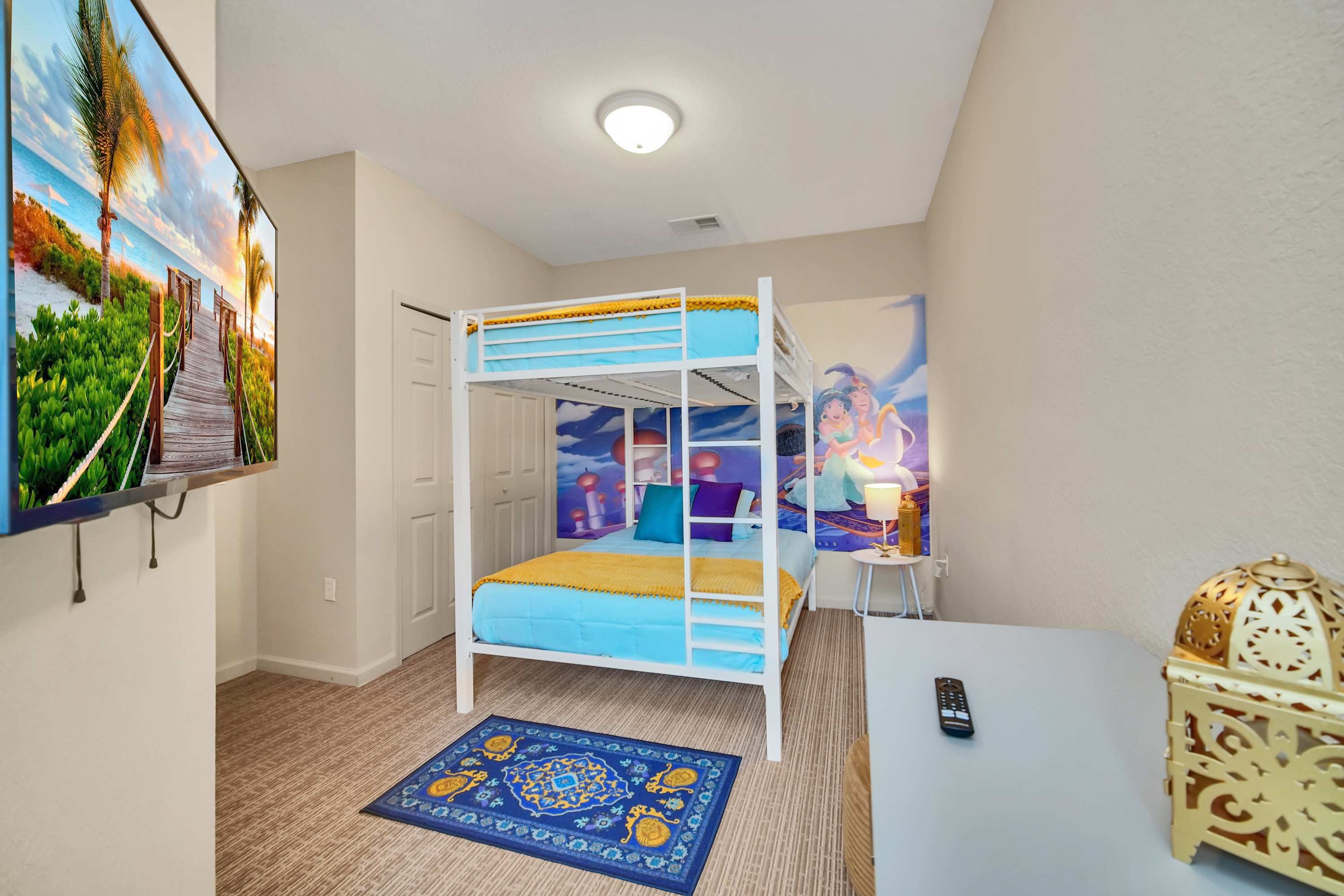 Themed kids' bedroom featuring a white metal bunk bed with blue and yellow bedding, a vibrant mural depicting Aladdin characters on the wall, and a wall-mounted TV.