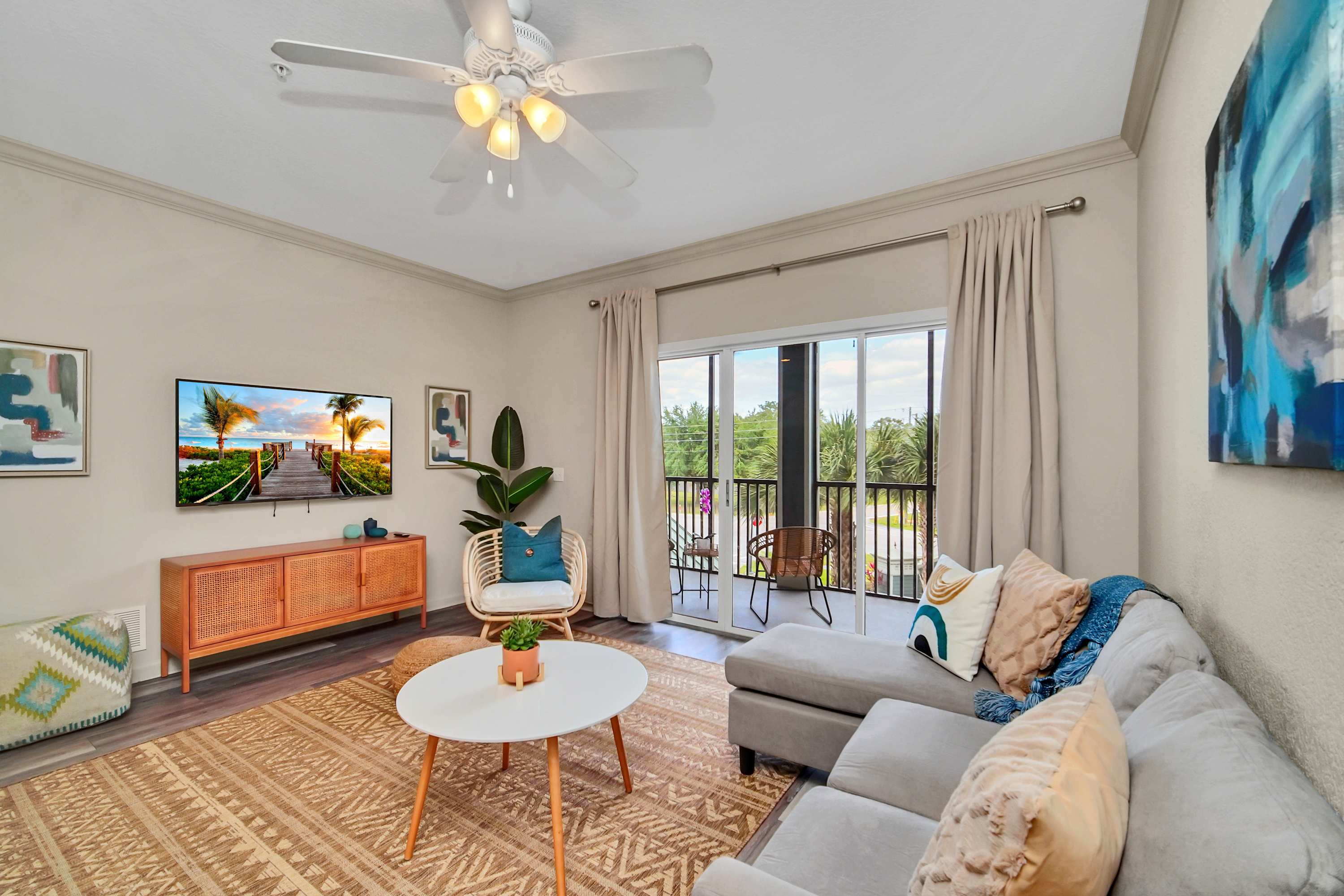 A bright living room with a light gray sectional sofa and a white round coffee table on a patterned jute rug. A wall-mounted TV is visible, and sliding glass doors open to a balcony with outdoor seating.