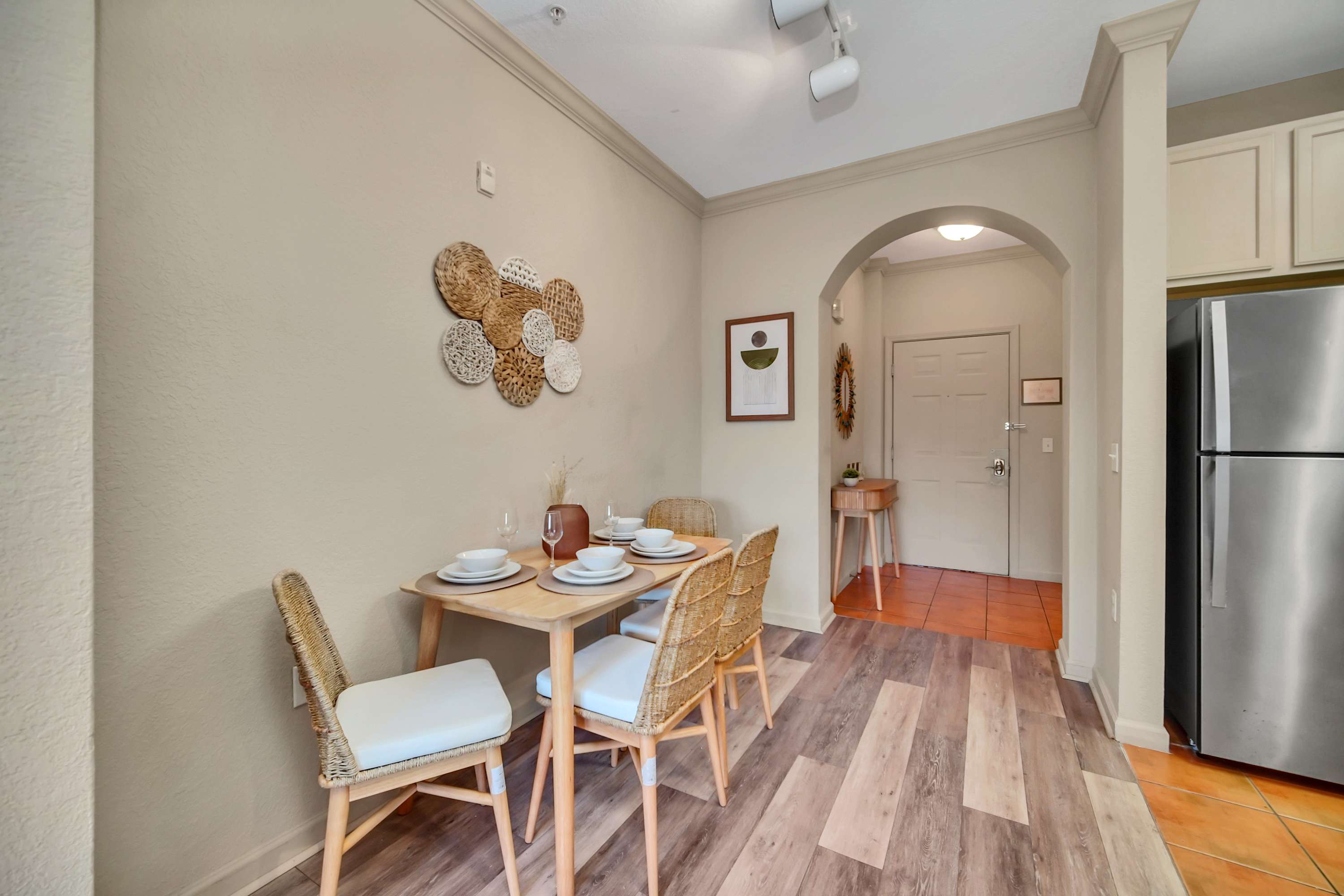 The dining area features a light wood table set for four with woven-back chairs, illuminated by track lighting, and decorated with circular woven wall art, leading to an arched entryway and a glimpse of the kitchen.