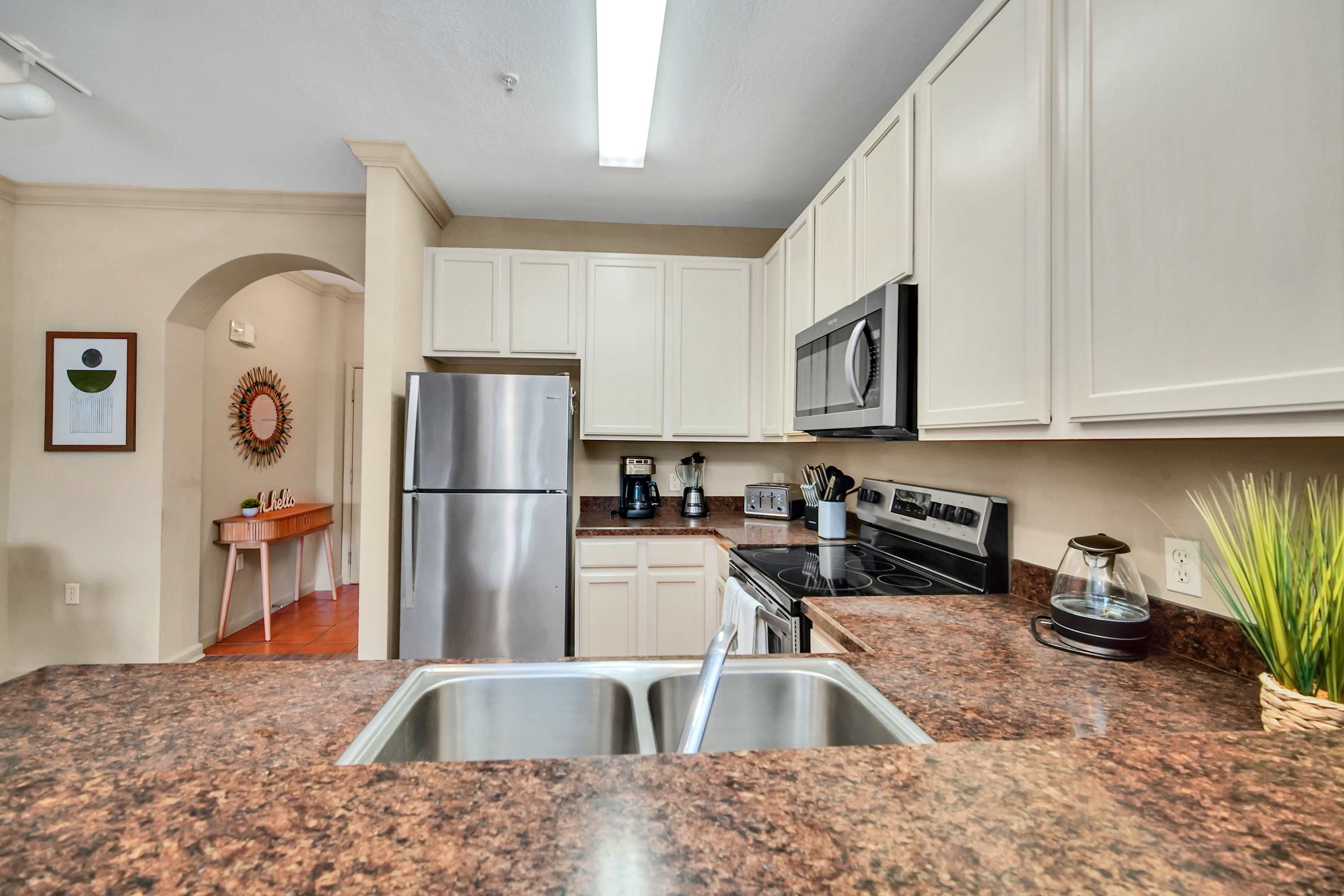 View from the kitchen sink area showing a double stainless steel sink, dark speckled countertops, and light cream cabinets, with a glimpse of the dining area and foyer beyond an arched opening.