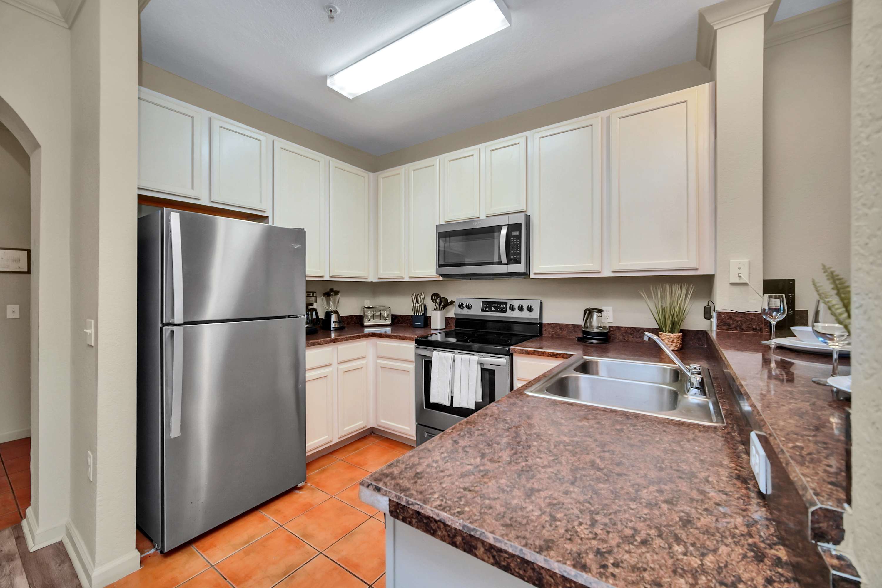 The kitchen features a double stainless steel sink set into dark speckled countertops, surrounded by light cream cabinets and stainless steel appliances, with terracotta-colored tile flooring.