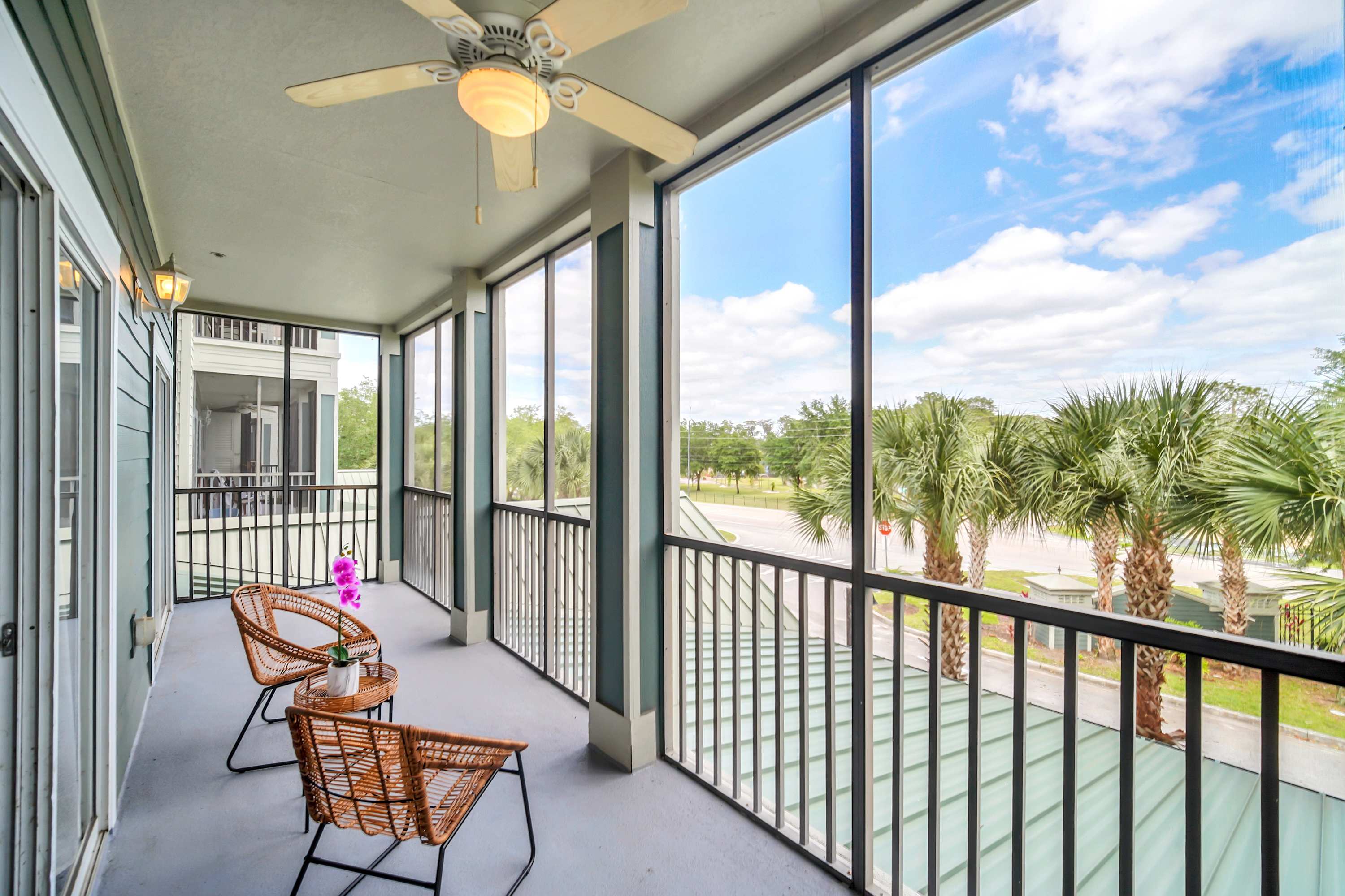 An expansive screened balcony with two rattan chairs and a small table, overlooking palm trees and a street view. The balcony features a ceiling fan and provides a bright outdoor living space.