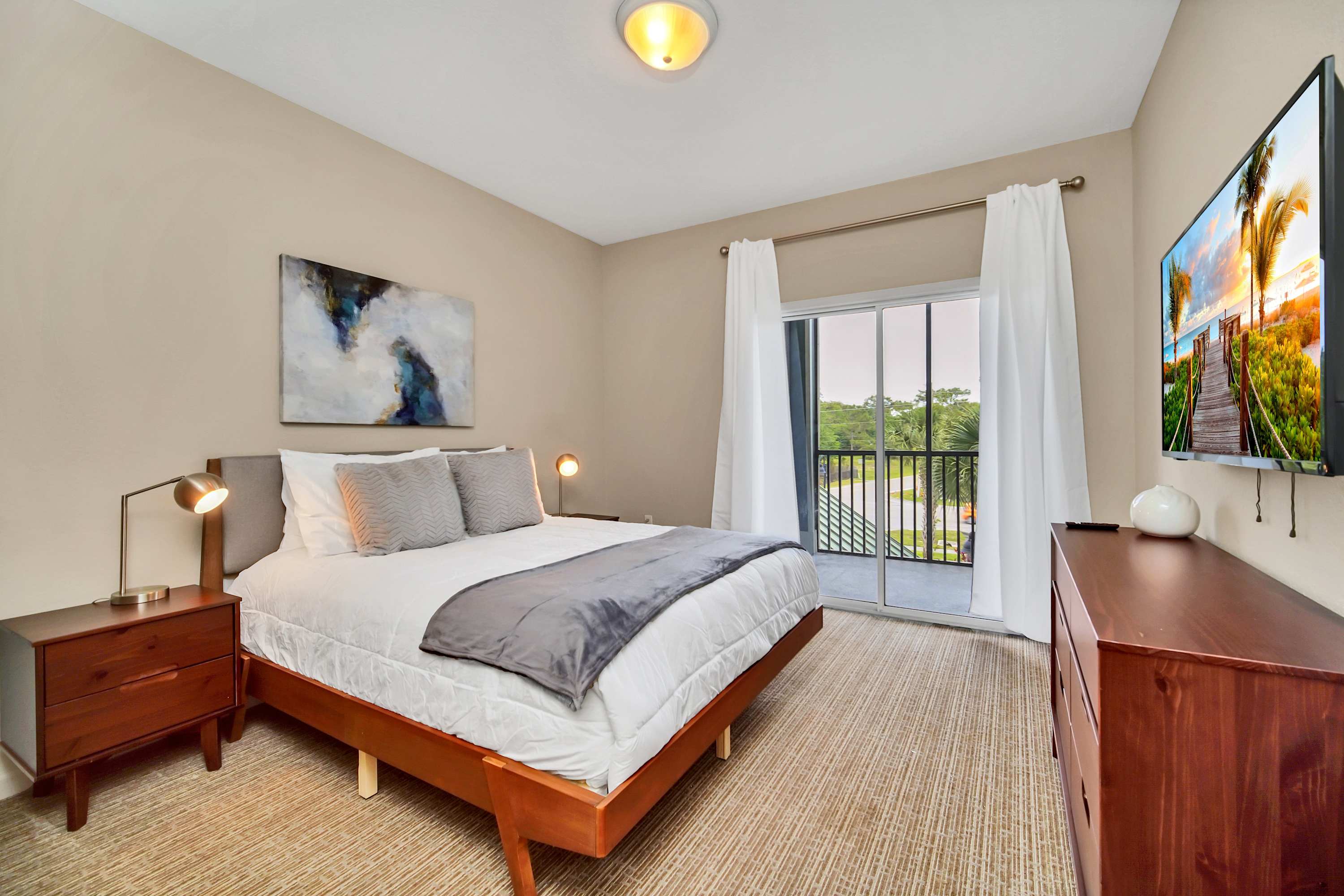 Bright bedroom featuring a queen-sized bed with a wooden frame, two nightstands with modern lamps, a wall-mounted TV, and a sliding glass door leading to a private balcony with outdoor views.