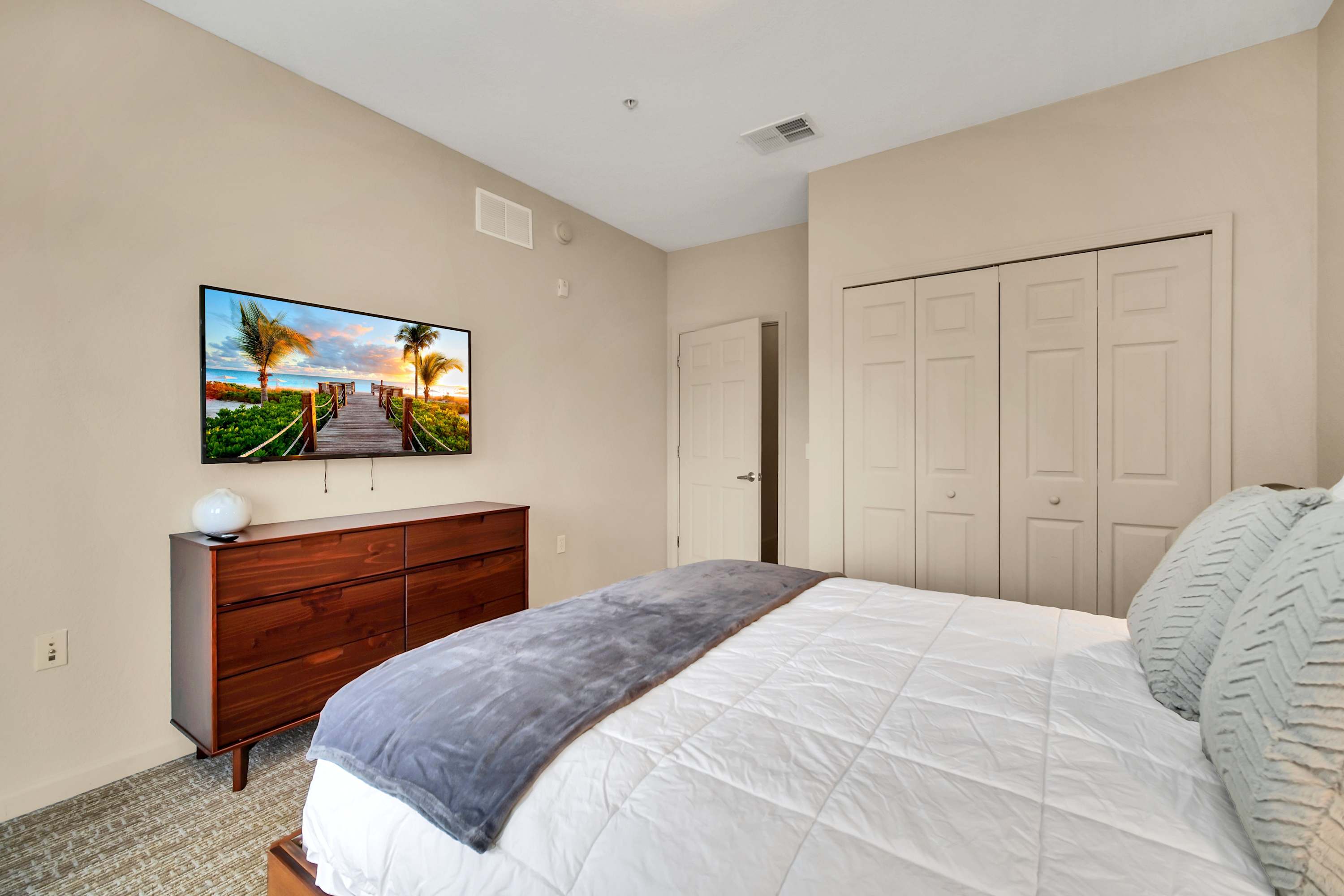 Another angle of the bedroom, showcasing a dark wood dresser with a wall-mounted TV above it, and a large closet with bi-fold doors, providing ample storage space.