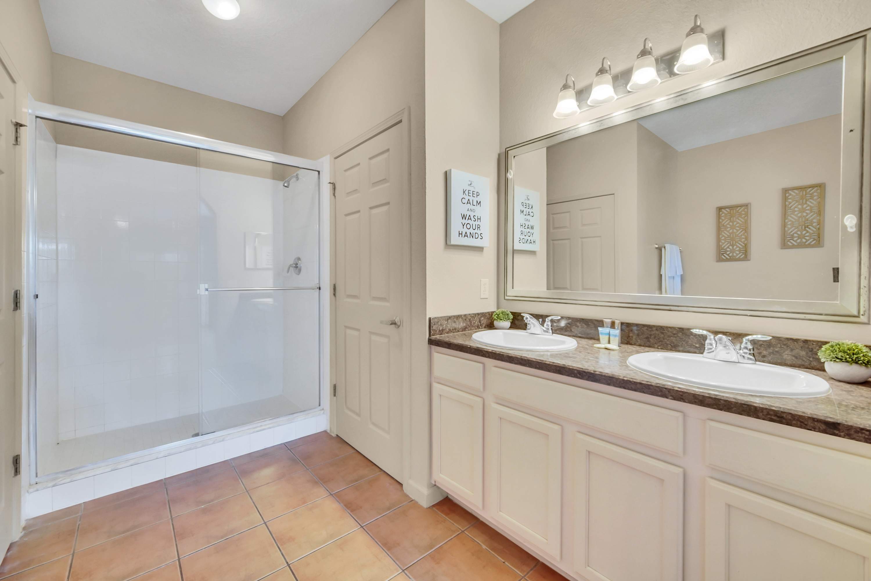 A bright bathroom features a double vanity with two sinks set into dark countertops, a large mirror, and a glass-enclosed walk-in shower, all with terracotta-colored tile flooring.