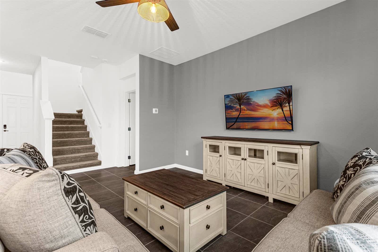 A ground-floor living room with dark tile flooring features two beige sofas, a light wood coffee table, and a wall-mounted TV against a dark gray accent wall. A carpeted staircase leads to the upper floor, and a white media console is positioned below the TV.