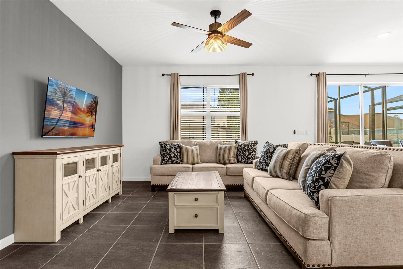 A spacious living room with dark tile flooring and white walls, featuring two large beige sofas with decorative pillows and a light wood coffee table with drawers. A white media console with a dark top holds a wall-mounted TV, and two windows with blinds and beige curtains provide natural light.