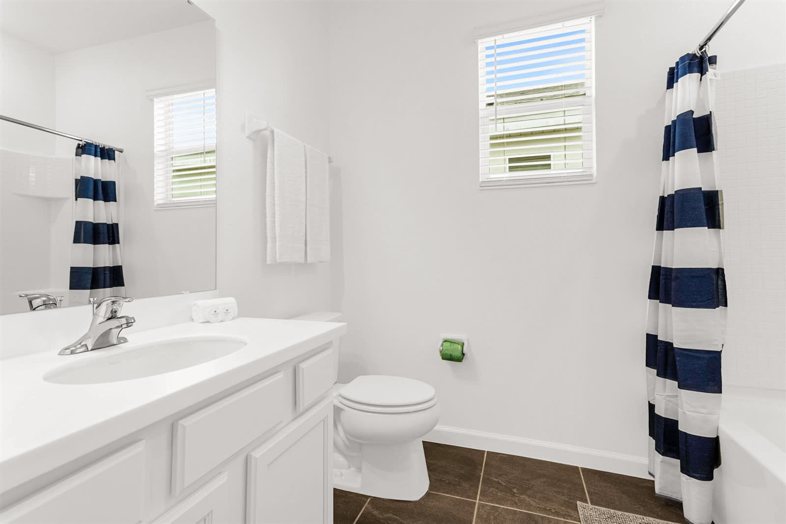 Bright bathroom with a white single vanity, toilet, and a shower-tub combo featuring a bold blue and white striped curtain, complemented by a window with blinds and dark tiled floors.