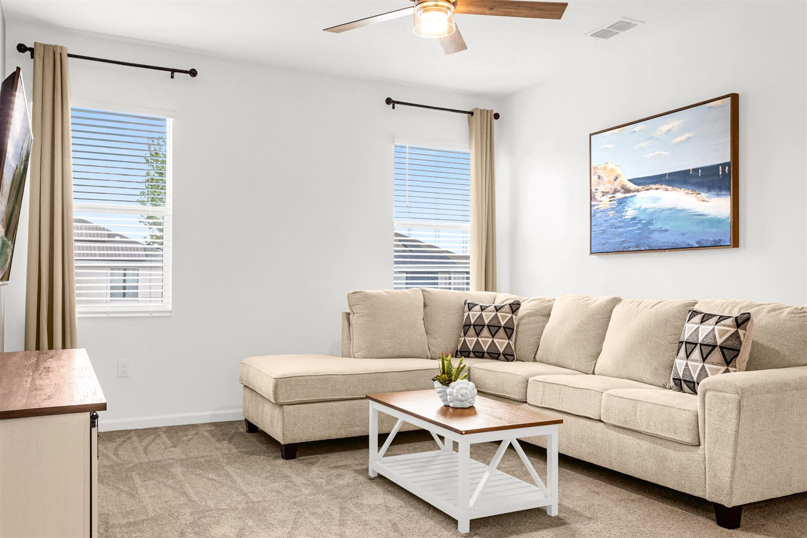 A cozy living room features a beige sectional sofa with patterned throw pillows, a wooden coffee table, and a wall-mounted TV. Two windows with blinds and curtains allow natural light, and a large painting of a beach scene hangs on the wall.