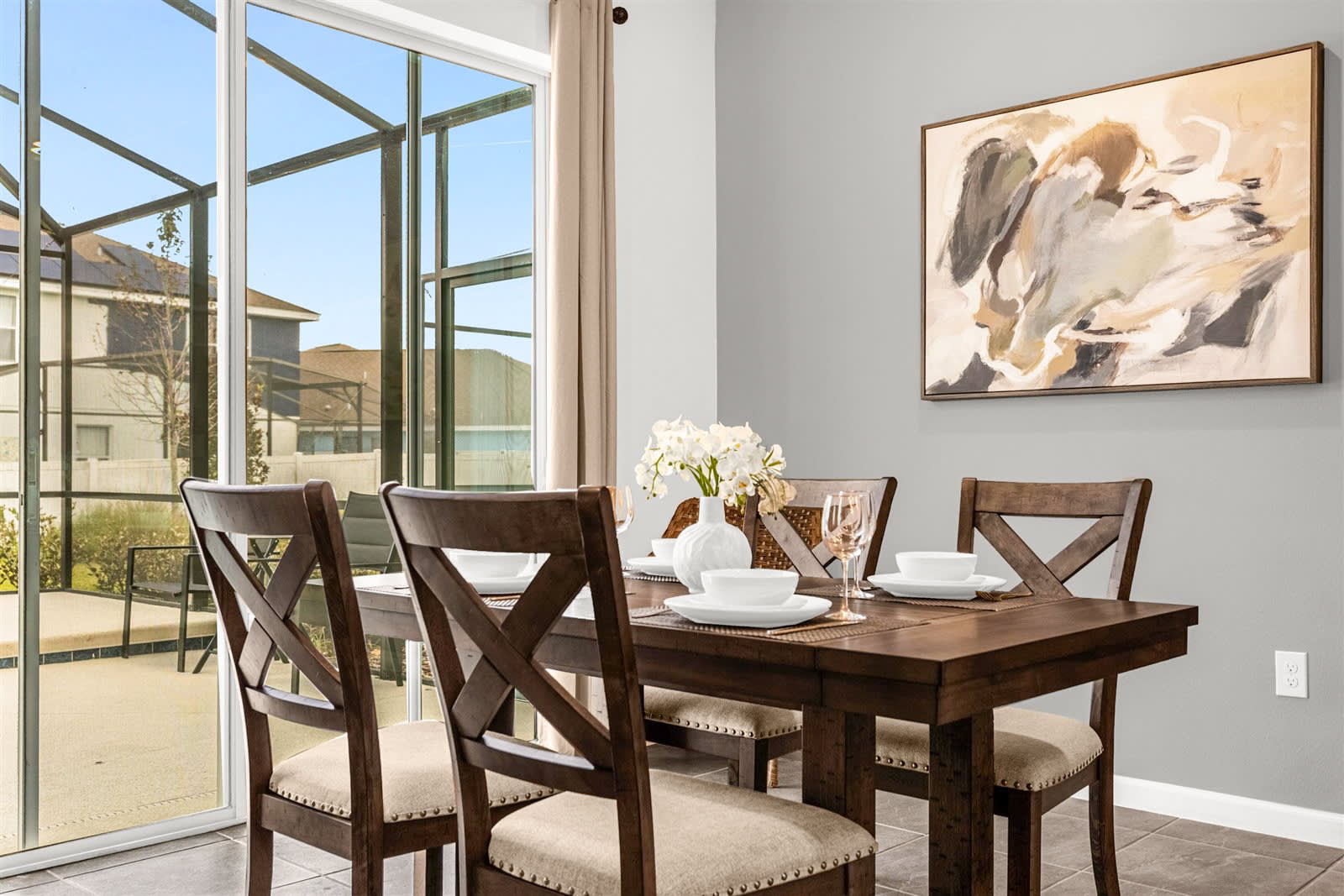 A modern dining area features a dark wood rectangular table set for four with white dishes and glasses, accompanied by four matching chairs with upholstered seats. A large abstract painting hangs on the light gray wall, and a sliding glass door offers a view of the screened pool area.