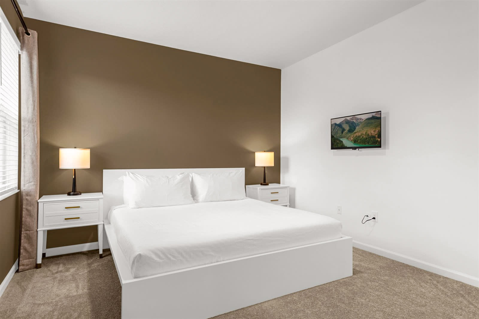 Modern bedroom with a white platform bed, two white nightstands with gold pulls, and a wall-mounted TV, set against a striking accent wall in a warm brown hue and light carpeted floors.