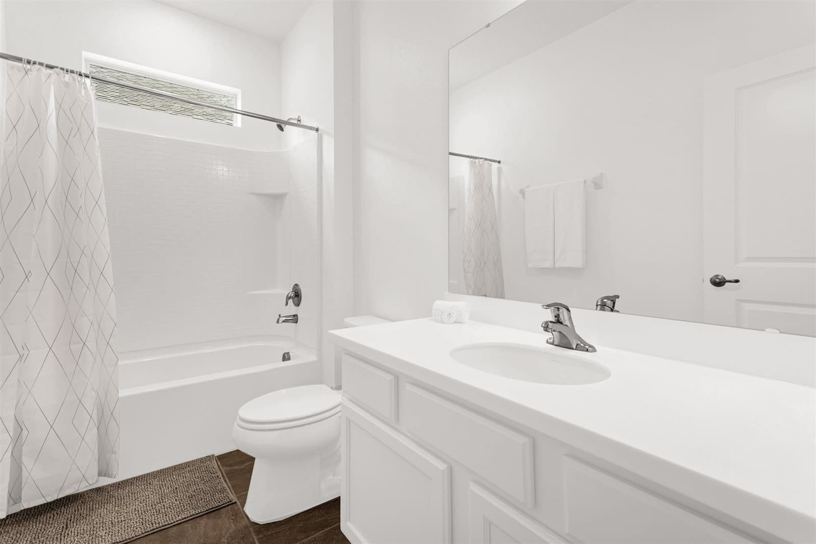 A bright, modern bathroom featuring a white vanity with a single sink, a large mirror, and a toilet. A shower/tub combo with white subway tiles and a patterned shower curtain is on the left.