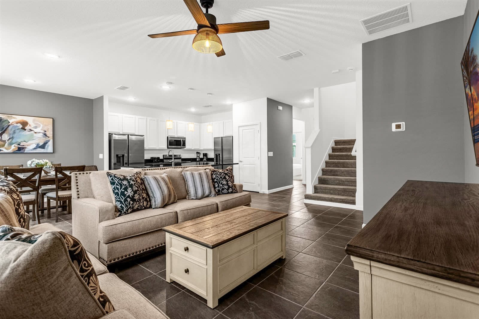 An open-concept living space with dark tile flooring connects the living room, dining area, and kitchen. The living room features two beige sofas and a coffee table, while the dining area has a dark wood table, and the kitchen boasts white cabinets and black countertops.