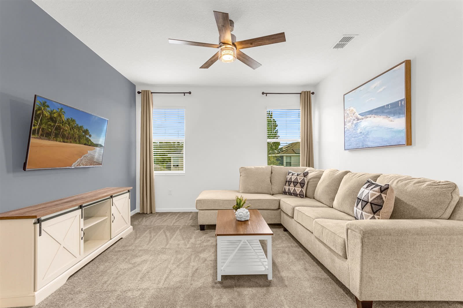 A cozy living room features a large L-shaped beige sectional sofa with patterned throw pillows, a light wood coffee table, and a wall-mounted TV displaying a beach scene. The room has light gray carpet, white walls, and a dark gray accent wall, with two windows covered by blinds and beige curtains.