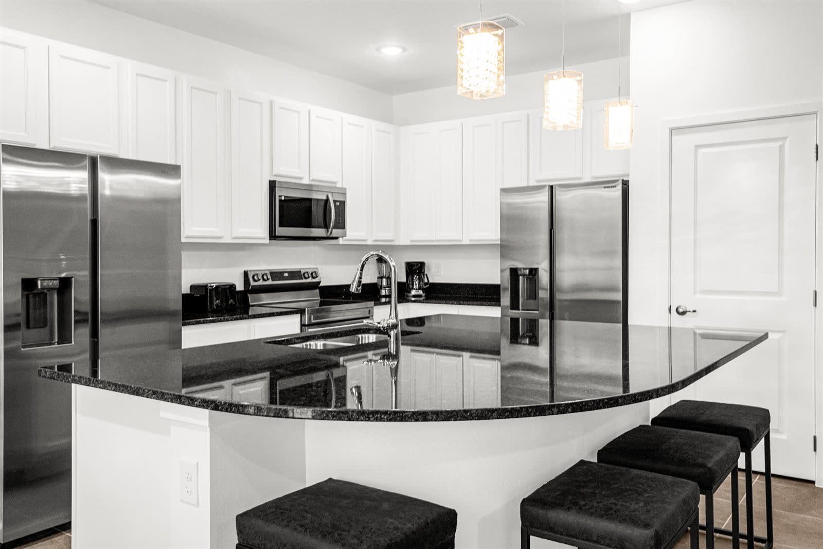 A modern kitchen features white shaker cabinets, black granite countertops, and stainless steel appliances, including a double-door refrigerator and a microwave. A large kitchen island with a sink and three bar stools is prominent in the foreground.